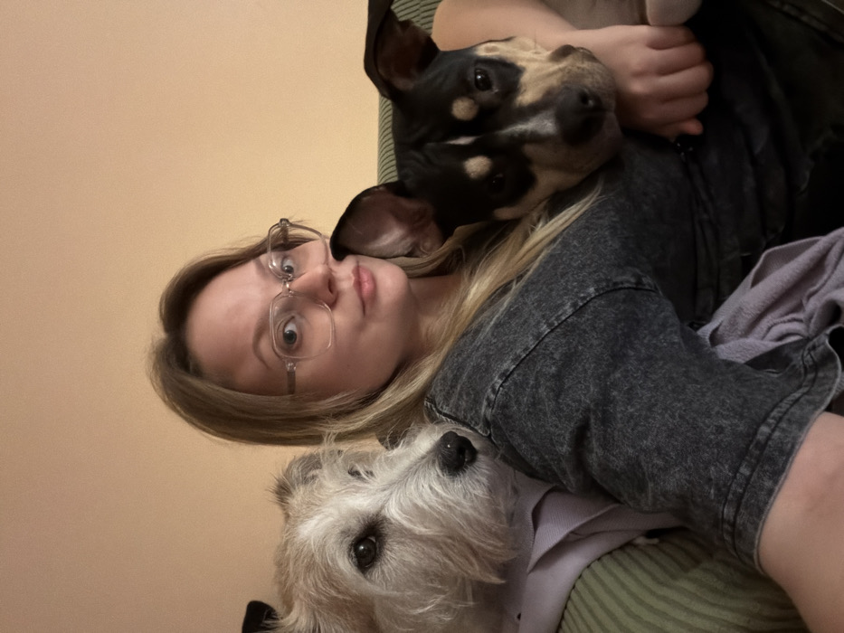 An indoor scene, specified as being in Tel-Aviv, Israel, shows a single adult female in close proximity to two dogs. The female, exhibiting light-colored hair and wearing clear-framed eyeglasses, is dressed in a dark grey, textured short-sleeved top. She maintains direct eye contact with the viewer. Positioned to her left is a light-furred, possibly terrier-type dog with dark eyes, resting its head on her left shoulder. A lavender-colored fabric is visible beneath this dog. To her right, a second dog with a black and tan coat and erect ears rests its head on her right shoulder, its body partially obscured. The subjects are seated on a piece of furniture upholstered with green and white striped fabric. The background consists of a plain, light orange-colored wall. The overall scene depicts a calm, domestic interaction.
