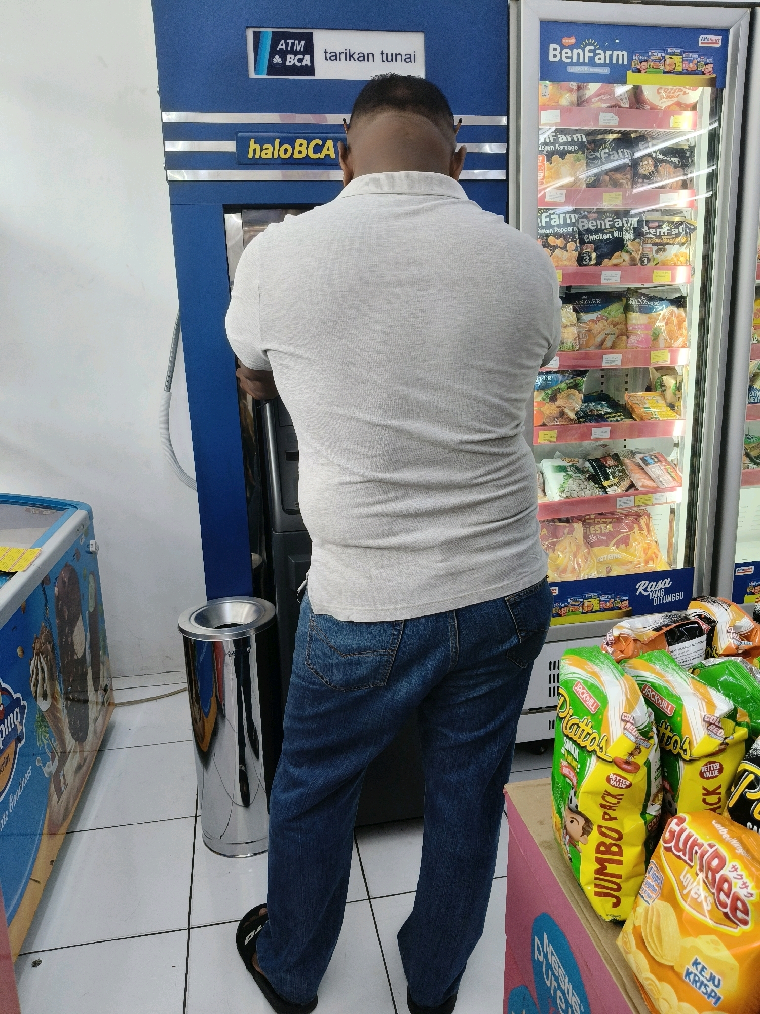 A man is standing in front of a blue ATM machine with "haloBCA" written on it. He is wearing a light gray polo shirt and blue jeans. The ATM is located inside what appears to be a convenience store, as evidenced by a refrigerated display case filled with frozen food products to the right of the ATM. This display case has branding for "BenFarm" and "Alfamart". To the left of the ATM, there is an ice cream freezer with colorful graphics. In the foreground, on a pink shelf, are several bags of snacks, including "Jumbo Pack", "Gurigee Layers", and "Keju Krispi". A silver trash can is positioned between the ATM and the ice cream freezer. The floor is tiled with white squares. The scene is consistent with a retail environment in South Tangerang, Indonesia, likely an Alfamart minimarket.