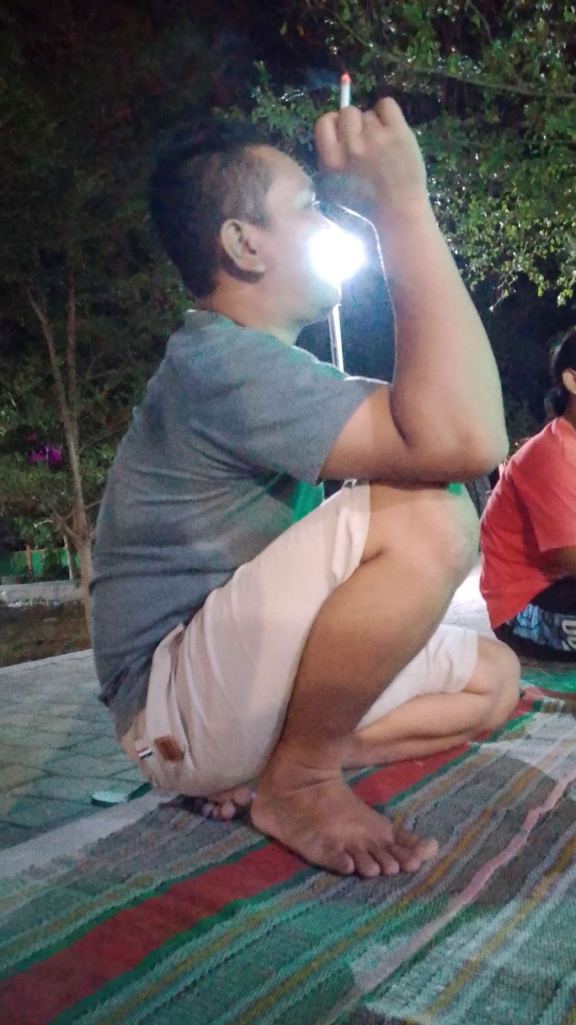 A man in a gray t-shirt and light-colored shorts is squatting on a striped mat outdoors at night, in what appears to be the city of Karanglangu, Indonesia. He is holding a lit cigarette to his mouth with his right hand, and his left arm is bent with his fist near his head. The background shows trees and foliage, and a bright light source is visible near his face, suggesting artificial lighting. Another person in a red shirt is partially visible in the background to the right. The ground appears to be paved.
