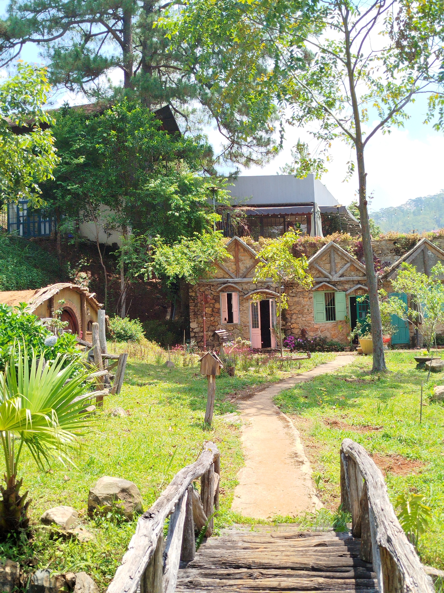 This vibrant outdoor scene captures a charming collection of rustic buildings nestled in a lush, green landscape, likely in Da Lat, Vietnam. In the foreground, a sturdy wooden bridge with weathered railings leads the viewer's eye along a winding dirt path that traverses a grassy area.
To the left, a unique, earthy structure resembling a Hobbit hole stands with a round wooden door, partially obscured by dense foliage. Further along the path, a series of quaint stone cottages with gabled roofs are visible. These whimsical dwellings feature colorful wooden doors and shutters in shades of pink, light green, and teal, adding a playful touch to their rustic stone facades. Purple flowering plants grow near their bases.
Above these cottages and further up the gentle slope, more modern structures with large windows and awnings are partially visible through the abundant trees and dense greenery. The entire scene is framed by various trees with rich green leaves, some appearing coniferous and others deciduous, under a bright, clear blue sky.
The weather is sunny, indicating late morning or early afternoon. A large, spiky palm-like plant is prominent in the left foreground, alongside natural rocks scattered in the grass. Small wooden birdhouse-like decorations are also interspersed throughout the garden. The overall atmosphere is peaceful and enchanting, suggesting a serene retreat or resort. No people are visible, and there is no discernible text.