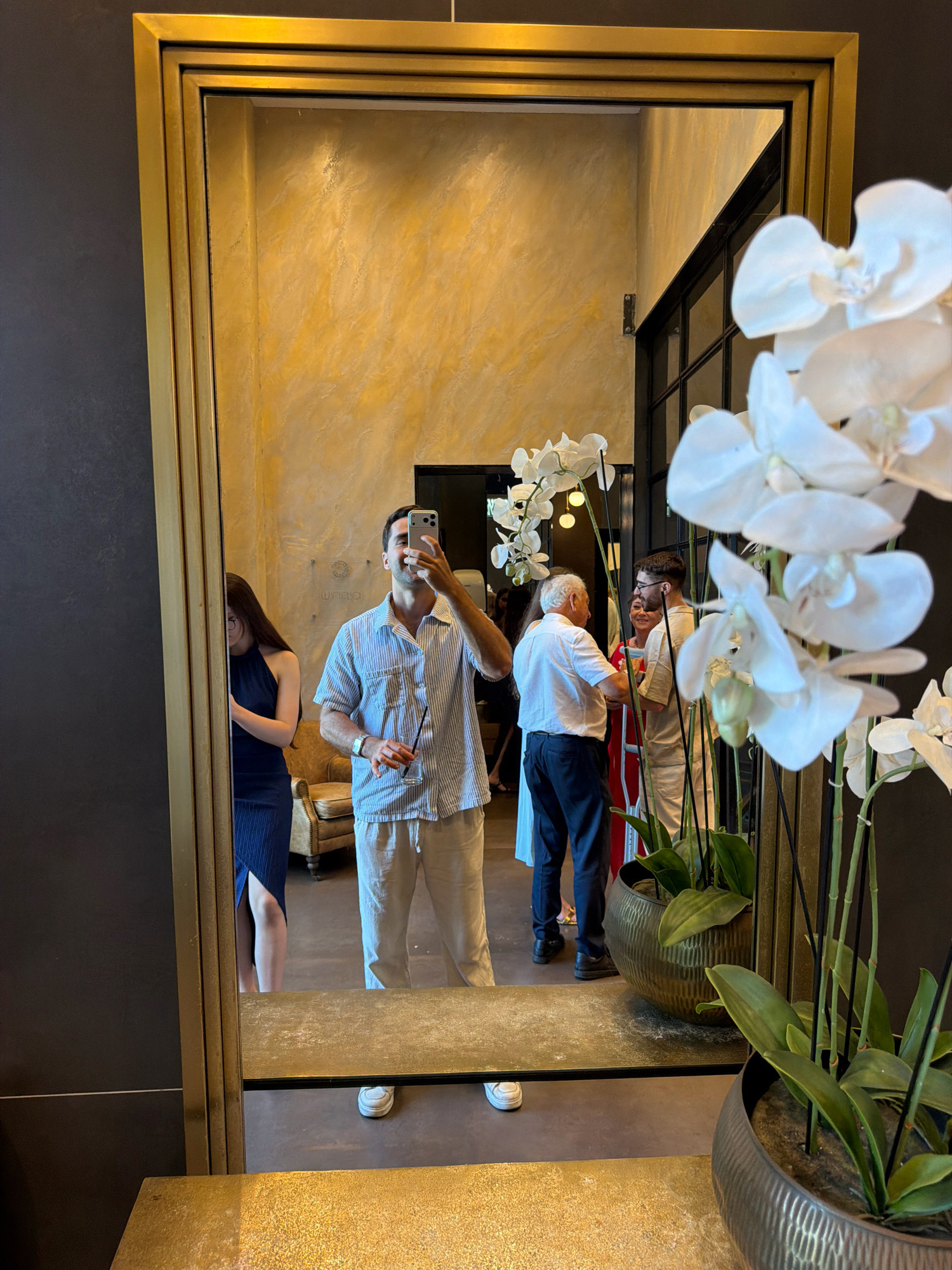In Kadima, Israel, a full-length mirror with a metallic gold frame reflects a group of people inside a well-lit interior space. In the foreground, a person is taking a selfie, holding a phone in their right hand and a glass with a straw in their left. They are wearing a blue and white striped short-sleeved shirt and light-colored pants. To the left of the selfie-taker, a woman in a dark blue dress is visible in the mirror, facing away and partially obscured. In the background of the reflection, other individuals are seen standing and conversing, with a plush armchair visible further back. Large white orchids in decorative pots are prominently displayed in the right side of the frame, partially obscuring the reflection in the mirror. The walls have a textured, light yellow finish, and a metal-framed window or partition is visible.