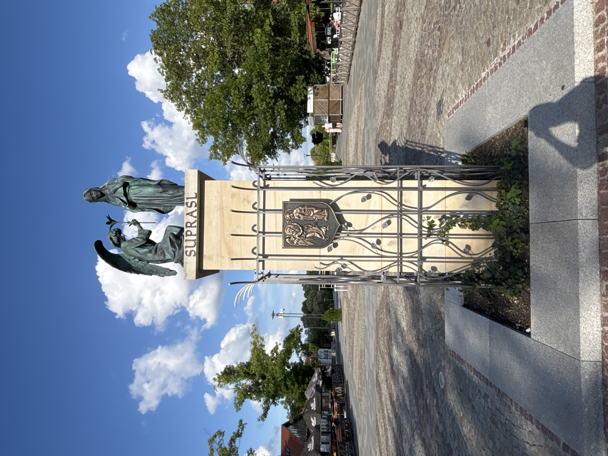 The image shows a bronze statue of an angel kneeling before a woman, with the word "SUPRASL" inscribed above them on a stone monument. The monument is surrounded by a decorative metal fence with a coat of arms featuring two winged figures. The scene is set in a public square paved with cobblestones, with trees and buildings in the background. The sky is blue with white clouds. There are some people visible in the distance, as well as outdoor seating areas with umbrellas. The statue appears to be a significant landmark in the town of Ogrodniczki, Poland, possibly commemorating a religious or historical event.
