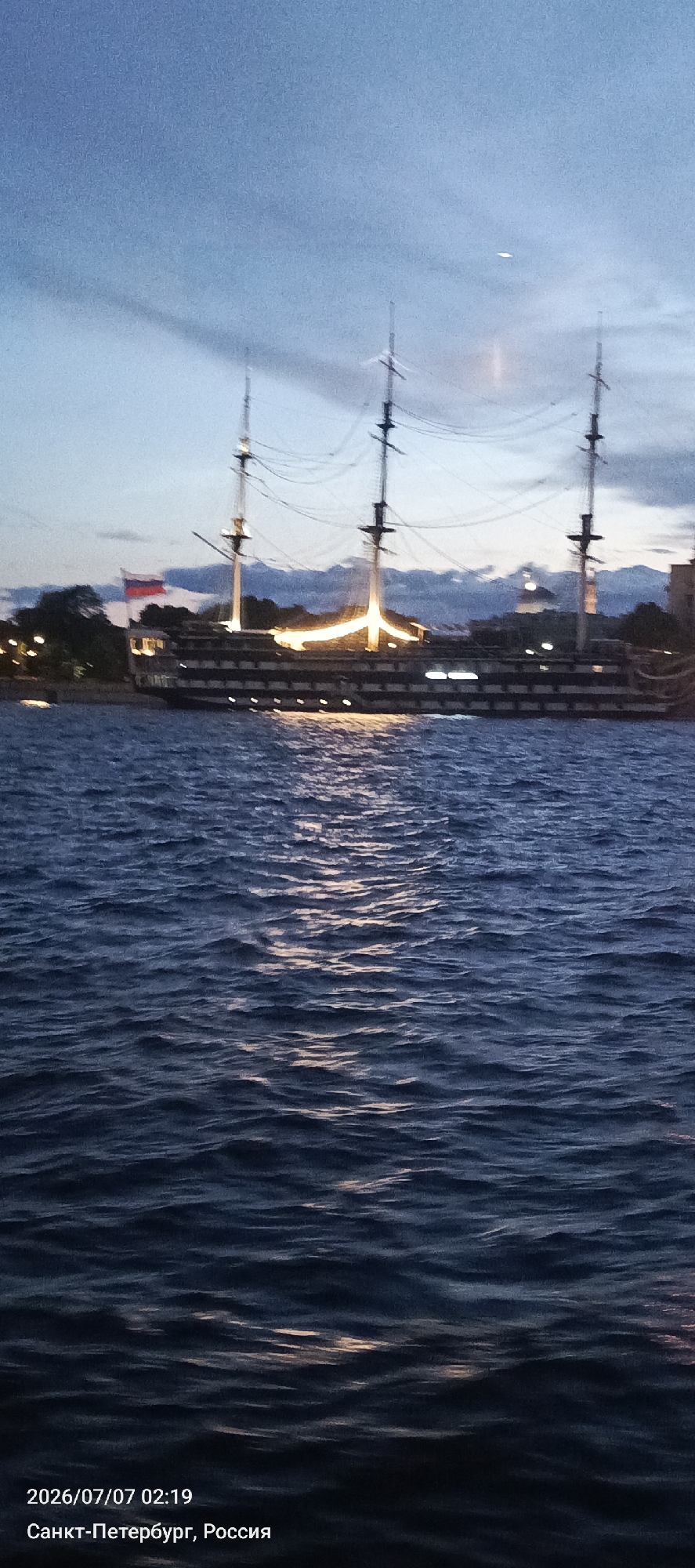 The image shows a wide, dark body of water with gentle waves under a twilight sky. In the background, a large, historic sailing ship is illuminated by warm lights along its deck. The ship has multiple masts with rigging. A Russian flag is visible on the ship. Trees and some lights from the shore can be seen behind the ship. The timestamp at the bottom indicates the photo was taken on July 7, 2026, at 02:19 in Saint Petersburg, Russia.