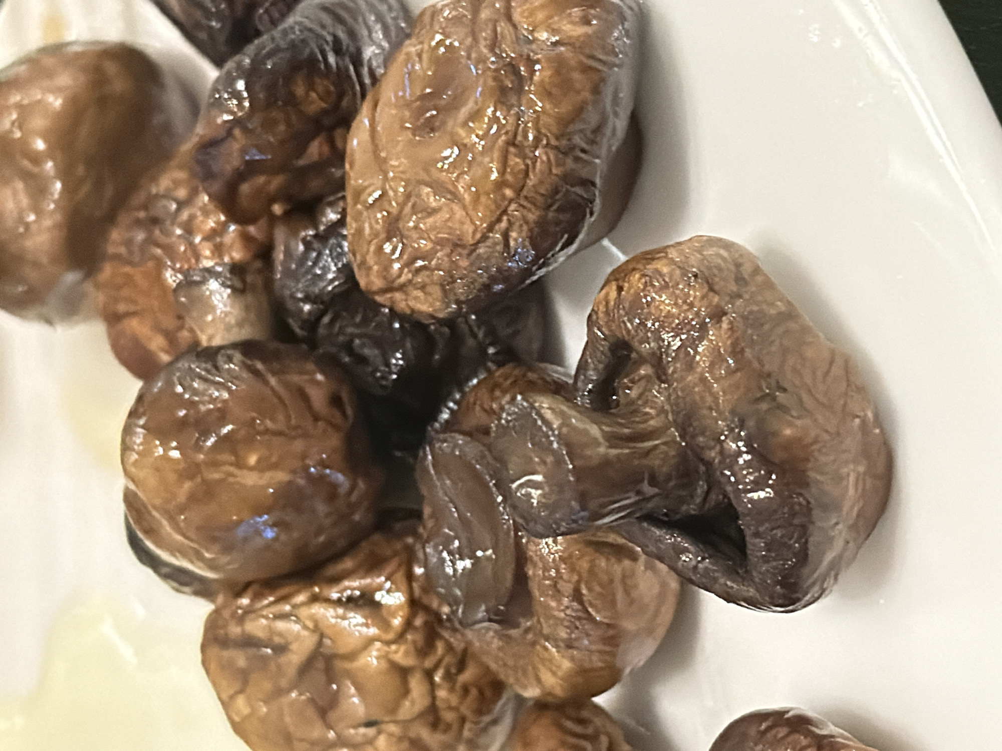 The image shows a close-up of a pile of cooked or marinated mushrooms on a white plate. The mushrooms are various shades of brown, some darker than others, and appear slightly glossy. The background is blurry, with a hint of what might be a dark green surface. There is no indication of the city of Moscow, Russia, or any people.
