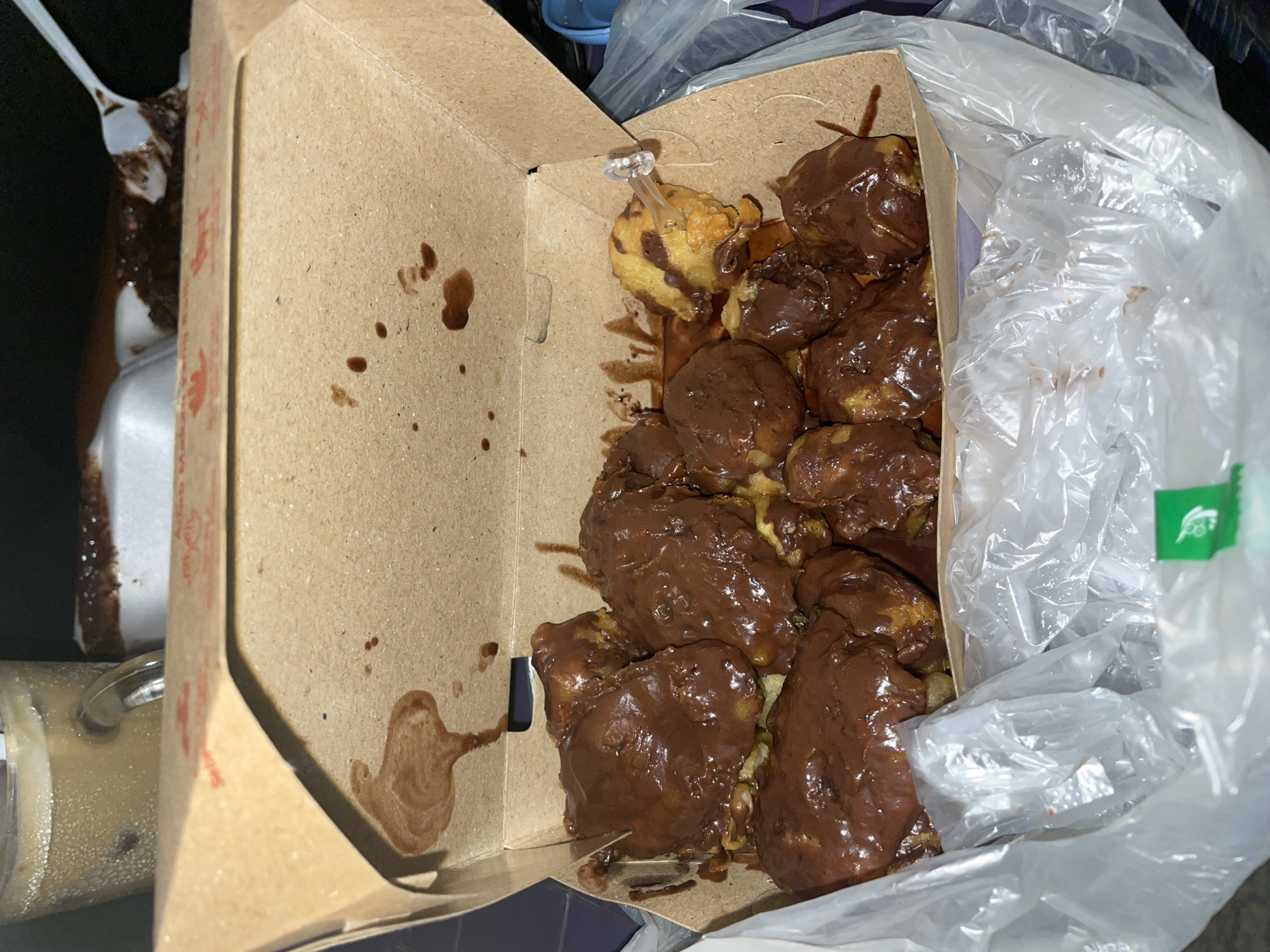 The image shows a cardboard box filled with chocolate-covered donut holes. The box is partially open, and a plastic bag is draped over it. A clear plastic utensil is visible near the box, along with a cup of coffee. The location is unknown.
