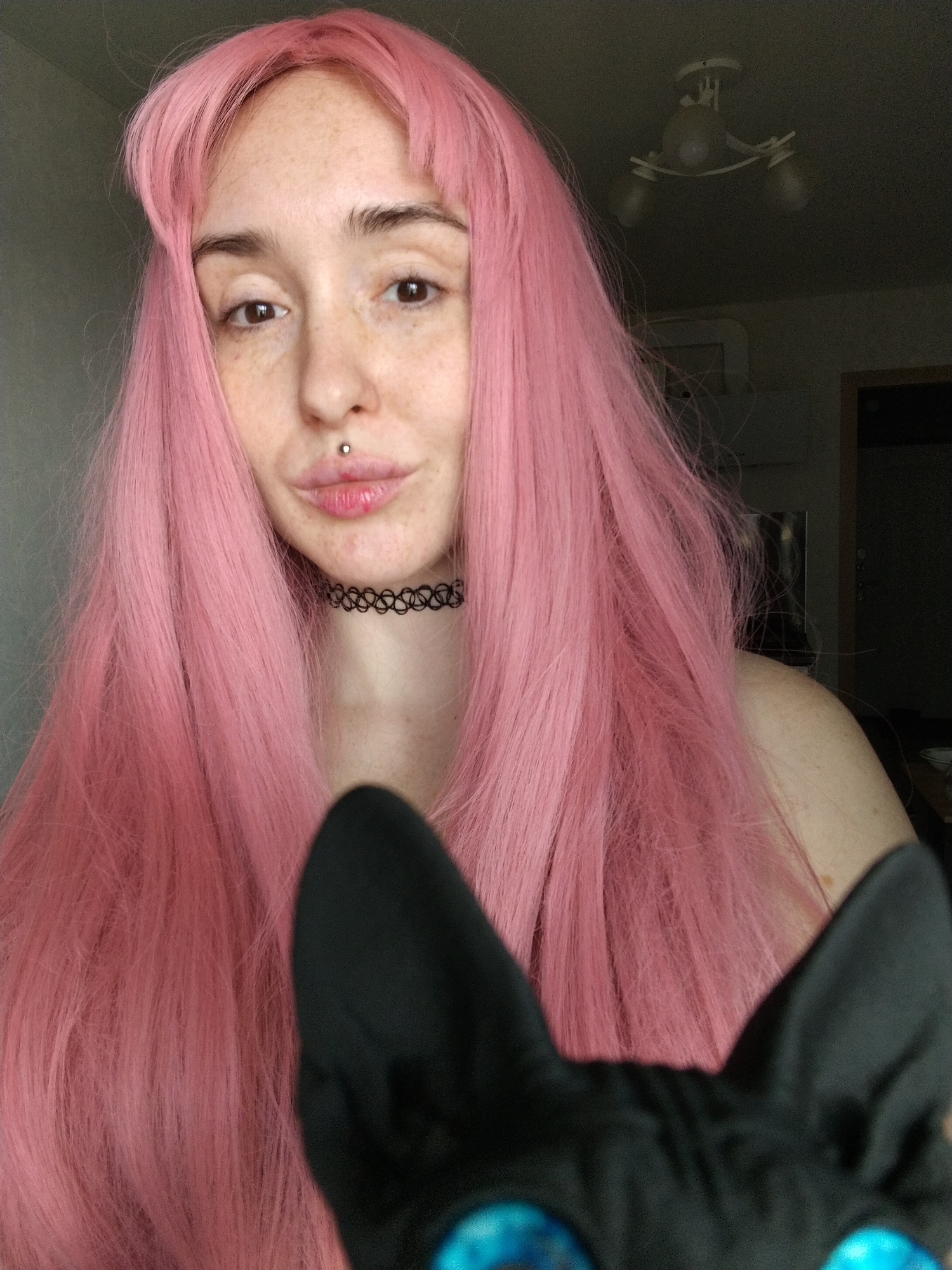 A fair-skinned individual with freckles and long, pink hair is the primary subject of the photograph. They have a labret piercing and are wearing a black choker necklace. The top of a black object, possibly a cat or a plush toy resembling a cat, is visible in the foreground at the bottom of the frame. The background is a room interior with a ceiling light fixture. There is no information in the image to indicate the location in the city of Izhevsk, Russia.