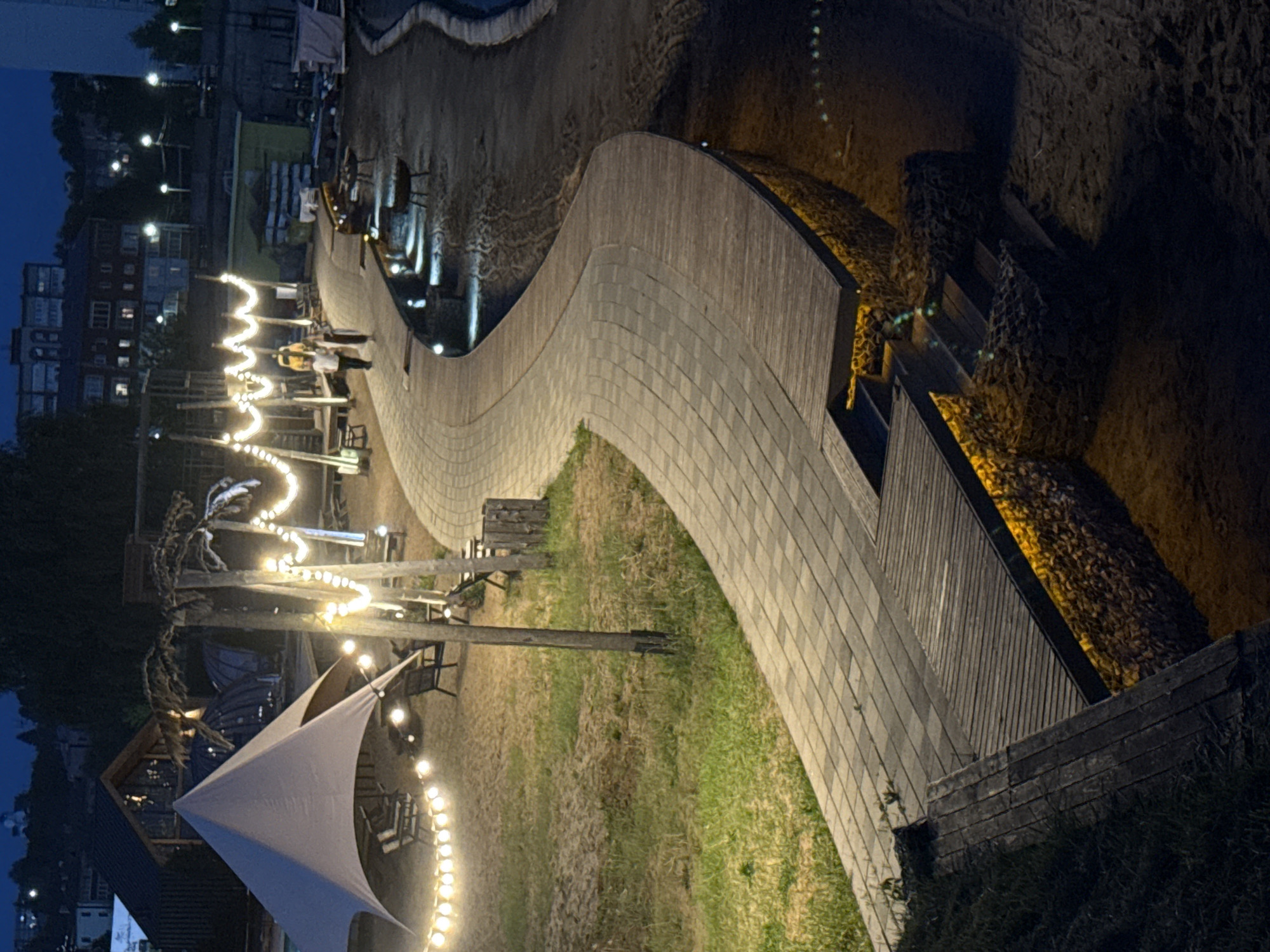 The image depicts a pedestrian area in Votkinsk, Russia, at night. A winding, elevated walkway constructed from light-colored wood bricks dominates the scene. The walkway features a wooden railing along its edge. Adjacent to the walkway is a grassy area with scattered vegetation. String lights are strung overhead, illuminating the area. Several wooden structures, including a sail-like canopy and timber posts supporting the string lights, are visible. In the background, a row of buildings and streetlights can be seen. The walkway descends via wooden steps at the right edge of the image. The lighting suggests that the image was taken during twilight or early evening.