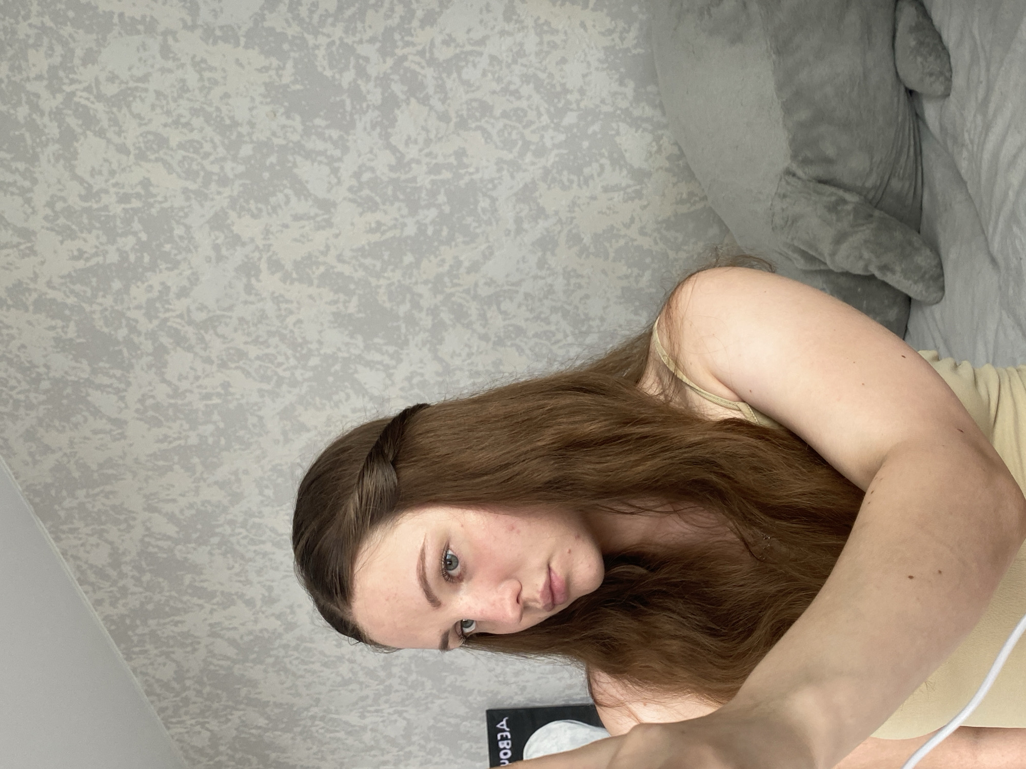 A young woman with long brown hair, some of which is braided and swept to the side, is shown in a close-up shot. She is looking away from the camera, towards her left, with a neutral expression. She is wearing a light-colored tank top. Her left arm and hand are visible in the foreground, and she appears to be holding a phone or camera. In the background, a gray wall with a textured pattern is visible. To the right of the woman, a large, gray stuffed animal resembling a manatee is resting on a surface, likely a bed. The overall lighting suggests an indoor setting. The image does not provide any specific information about the location in the city of Izhevsk, Russia.