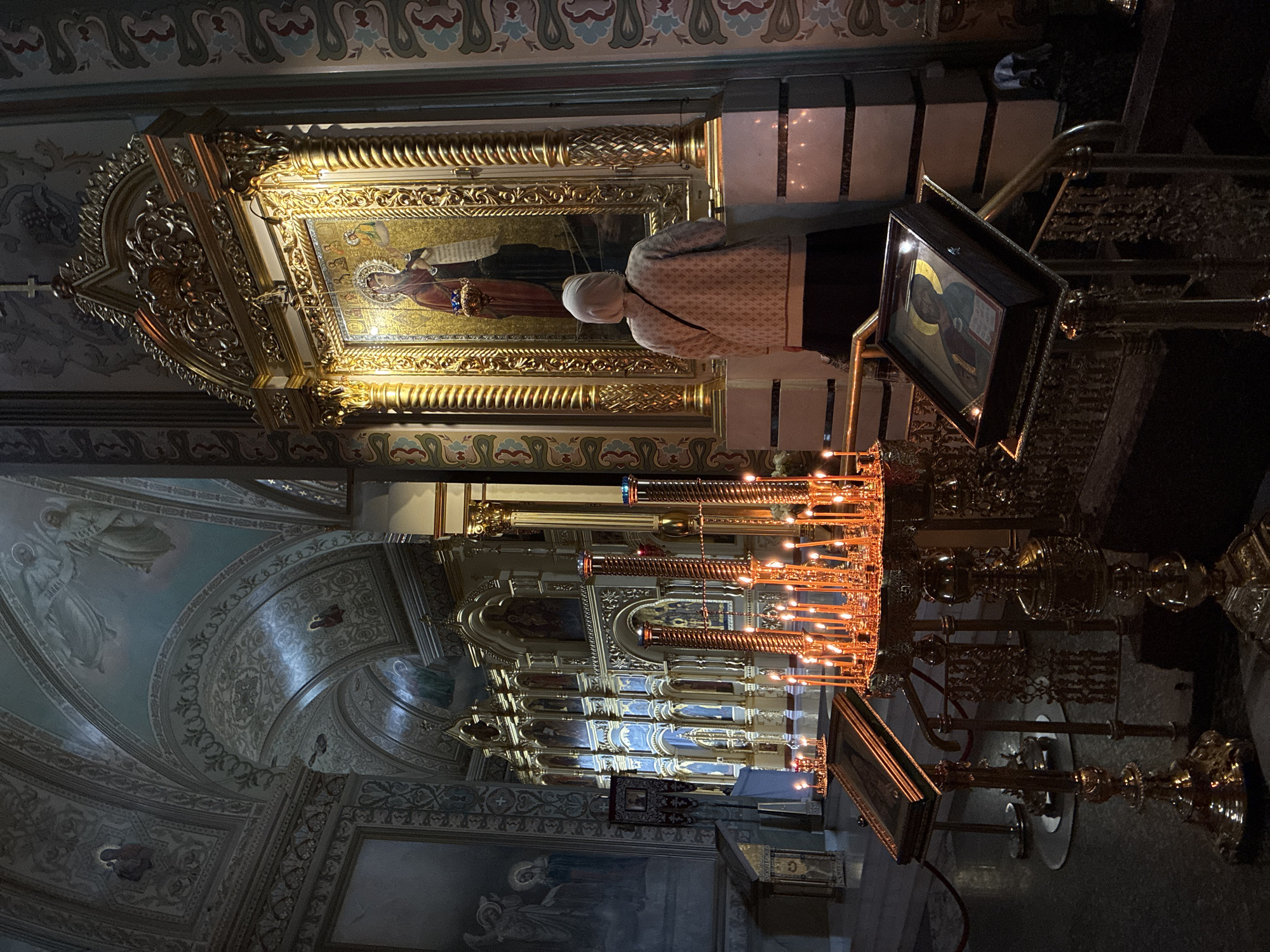 The image captures the interior of the Боголюбово Monastery in Russia, a site marked by elaborate religious ornamentation. A woman with a head covering stands near a golden shrine or altar, which is adorned with a painting of a religious figure. In front of her, a dense array of lit candles adds warmth to the scene. An ornate golden frame displays a smaller icon of a saint. The interior features painted ceilings and walls, further emphasizing the spiritual nature of the location. The lighting is subdued, with the candles providing the primary illumination, enhancing the atmosphere of reverence.