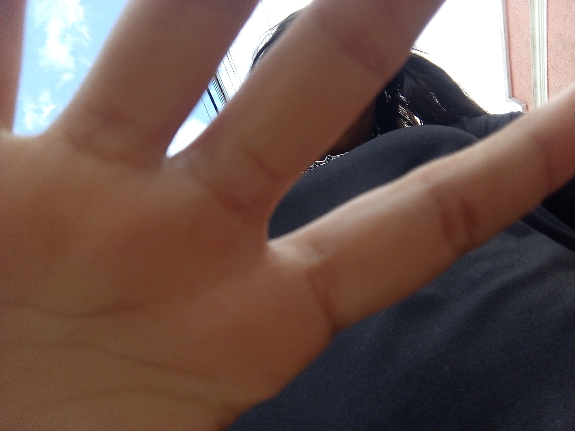 The image is a low-angle, close-up shot dominated by an outstretched human hand. The fingers are blurred due to proximity, obscuring much of the background. Behind the hand, a person with dark hair and wearing a dark top is partially visible. A chain is visible around the person's neck. In the top left corner, patches of blue sky with clouds are discernible. A corner of a building with pink siding and white trim can be seen in the background. The specific location within the city of Juan Griego, Venezuela cannot be determined from the image.
