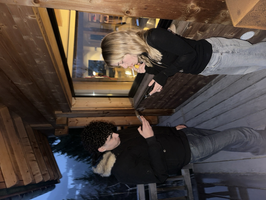 On an exterior wooden balcony or deck in La Clusaz, France, two individuals are visible during twilight or early evening. The male on the left, with dark curly hair, wears a black winter jacket featuring a light-colored fur-lined hood, and dark blue jeans. He is looking down at a smartphone held in both hands. The female on the right, with blonde hair, wears a black long-sleeved top and light-wash jeans. She holds a smartphone in her left hand and a lit cigarette in her right hand, positioned near her mouth. Both individuals are focused on their respective devices. The balcony features wooden overhead beams and plank flooring. To the right, a large rectangular window reveals a warmly lit interior space with visible furniture and shelves. Beyond the balcony, a snowy, forested mountain landscape is discernible under dim natural light.