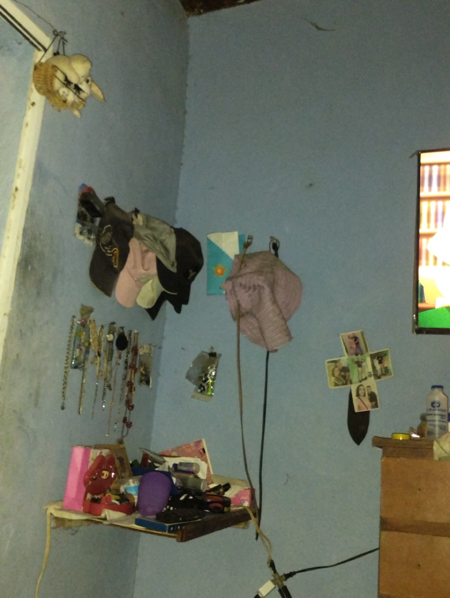 The image depicts a cluttered interior space, likely a bedroom or living area, in a residence located in La Guardia, Venezuela. The walls are painted a pale blue and appear somewhat worn.
Dominating the upper left of the frame is a decorative item, possibly a stuffed animal or a craft, hanging from a hook. It appears to be a white, possibly bunny-like figure with large ears, placed inside a woven basket.
Below this, to the left, is a shelf that holds various items. A pink rectangular object is on the far left, followed by a red object with visible eyes and a mouth, resembling a cartoon character. Behind these, other assorted objects and papers are visible, some appearing to be books or stationery. A purple object, perhaps a hat or a cushion, is also on the shelf.
The wall to the right of the shelf displays a collection of hanging items. Several necklaces and watches are suspended from hooks, creating a display of personal accessories. Further to the right, a pink and white striped fabric item, possibly a hat or a scarf, hangs from a hook.
On the wall, further to the right, are a few small framed photographs arranged in a cross shape. Below these photographs, a wooden dresser or cabinet is partially visible on the right side of the image. On top of this dresser, a blue plastic bottle with a white cap is present, along with some other small objects.
A television screen is visible in the far right background, displaying a colorful, abstract image. The overall impression is one of a lived-in space with personal belongings accumulated over time, reflecting a modest living situation in La Guardia.
