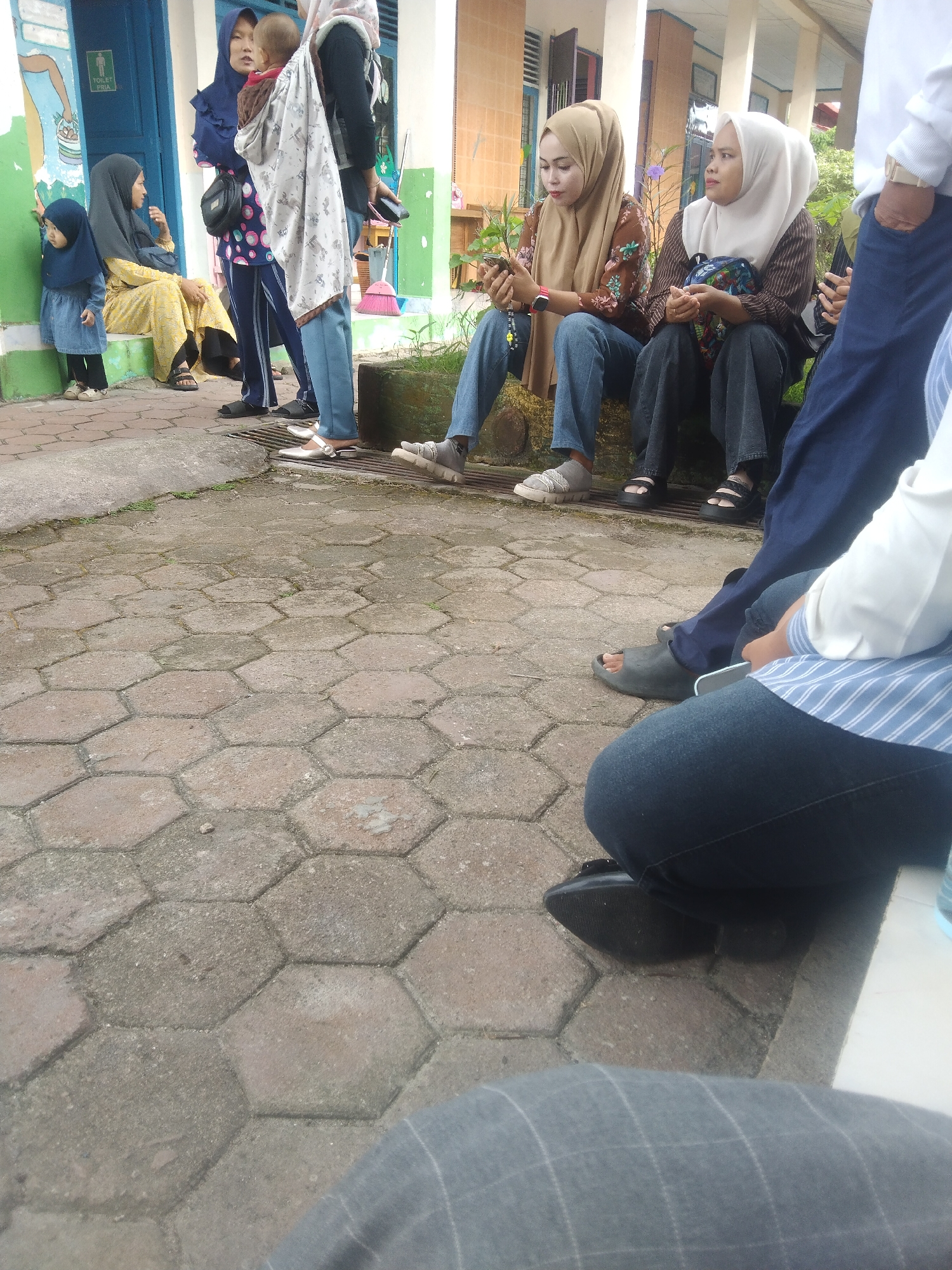 The image depicts an outdoor scene with several individuals, predominantly women, gathered near a building. The ground is paved with hexagonal concrete blocks. On the left, a young child wearing a blue headscarf and dress stands on green painted steps. Beside her, an adult woman in a yellow patterned dress and dark headscarf is seated. Further back, two women in dark headscarves and one in a patterned outfit stand near a blue door with a "TOILET PRIA" (Men's Toilet) sign. A woman in a light-colored headscarf is also present, holding a baby wrapped in a patterned cloth. In the foreground and mid-ground, several individuals are seated or standing. Two women in hijabs are seated on a raised surface, one holding a smartphone and the other looking away. Their attire includes patterned shirts and jeans. Other individuals' legs and feet are visible, wearing sandals, flip-flops, and sneakers. The building in the background appears to be a school or public facility, with a light-colored facade and windows. The scene suggests a casual gathering or waiting area in an urban or semi-urban environment. While the specific location of Siak Sri Indrapura, Indonesia is mentioned, there are no distinct visual cues within the image that definitively identify it as such.