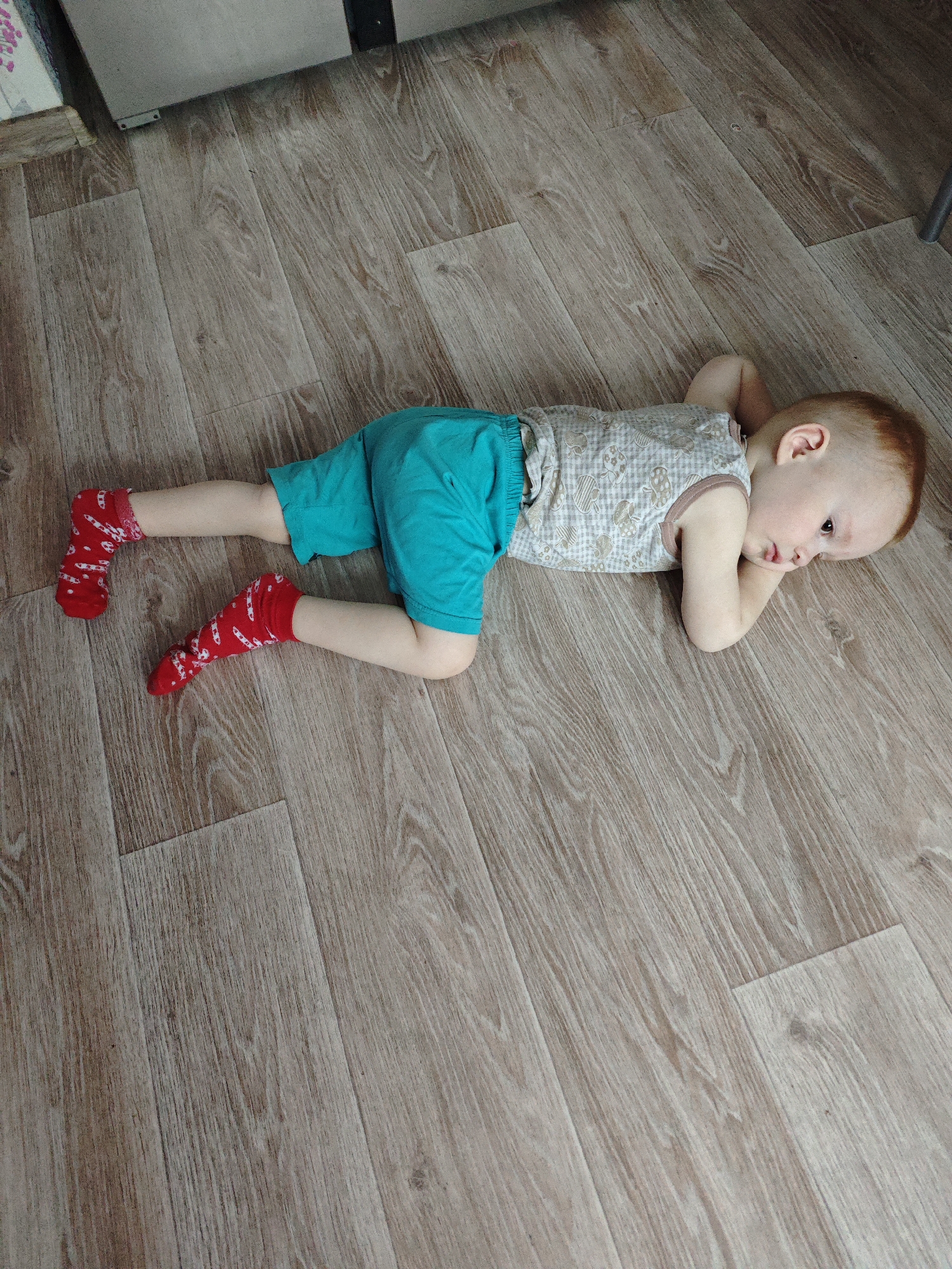 A young child lies prone on a wood-grain floor. The child wears a beige patterned tank top, teal shorts, and red socks with white designs. The child's arms are bent with hands near the head, and the child looks to the right. The floor appears to be made of planks. The location is Markovskii, Russia.