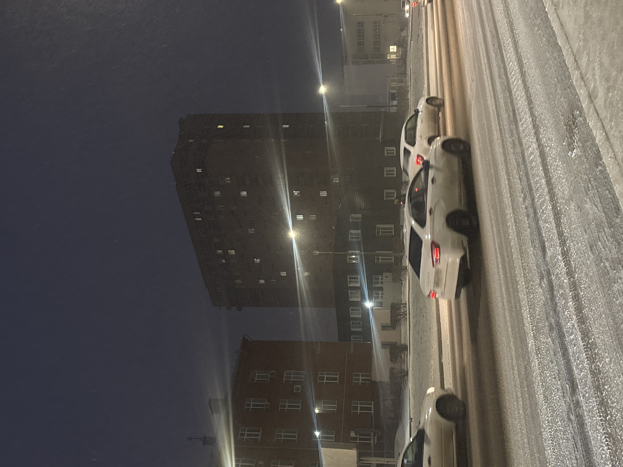 The image shows a night view in Perm, Russia, during snowy weather. Several cars are parked along a snow-covered street. A tall, dark apartment building dominates the background, with a shorter brick building adjacent to it. Streetlights illuminate the scene.