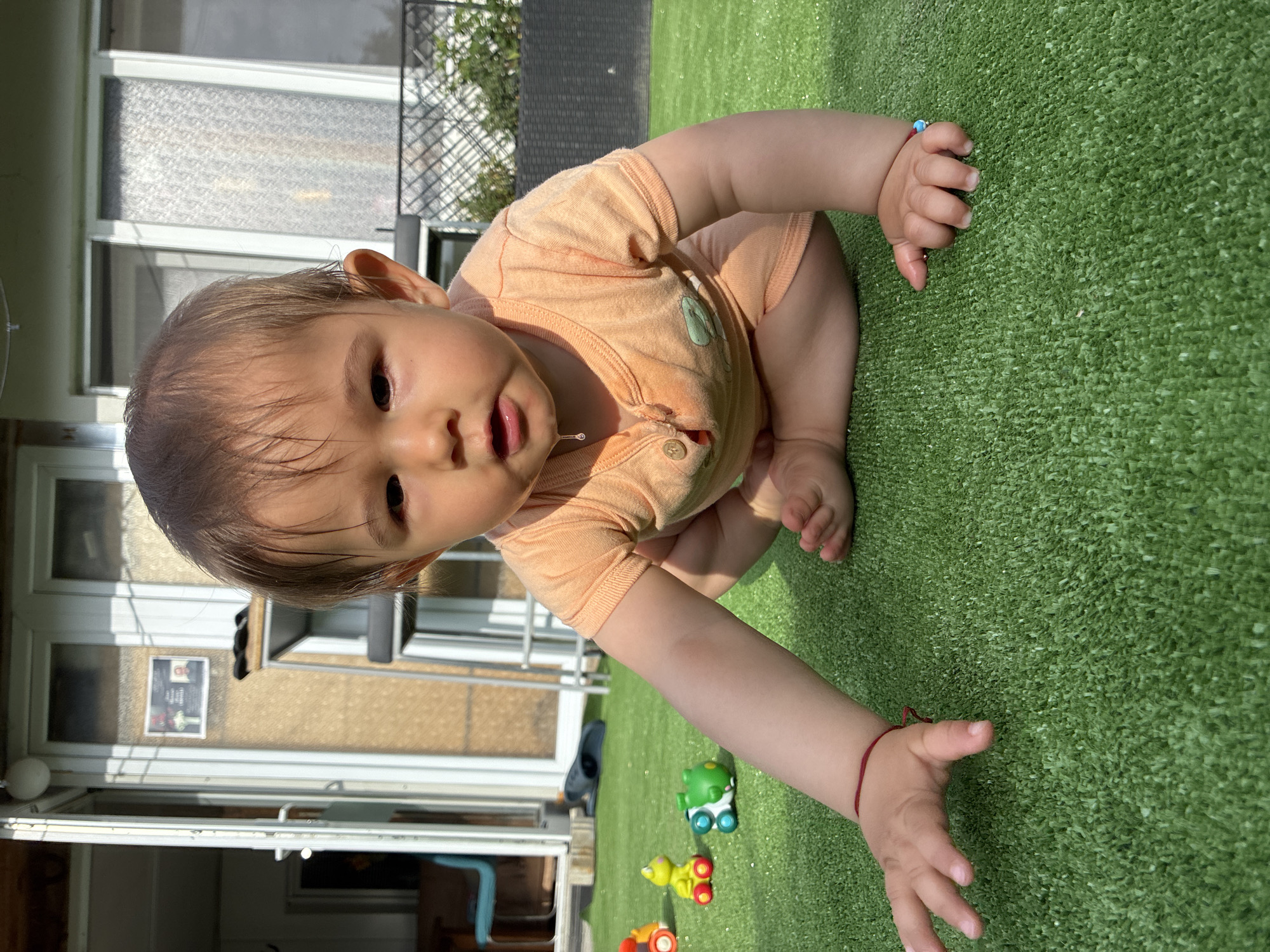 The image features a baby or toddler on a bright green artificial turf, likely an outdoor balcony or patio in Varna, Bulgaria. The infant, with brown hair, is in a low squatting or crawling position, looking directly at the camera with a curious expression. A drop of drool is visible on their chin. They are wearing a light orange romper with a subtle, light-colored emblem on the chest. A simple red string bracelet adorns the baby's right wrist, and a blue and red beaded bracelet is on the left.
Three small, colorful toy vehicles – green, yellow, and orange – are scattered on the artificial grass near the baby's outstretched right hand. In the background, part of a building with large glass doors or windows is visible, indicating an indoor space adjacent to the outdoor area. Through one of the glass panels, a blurry poster or sign with indecipherable text can be seen on the floor inside.
The scene is brightly lit by natural sunlight, suggesting a clear, sunny day, likely during the late morning or early afternoon, with visible shadows cast. The atmosphere is joyful and innocent, capturing a moment of a child's outdoor play.