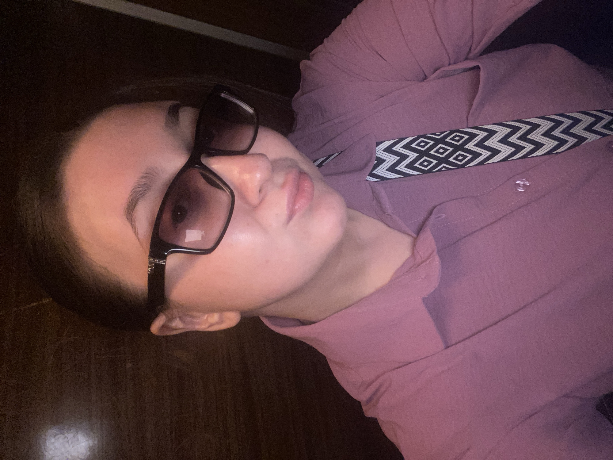 This is a close-up, rotated portrait image featuring a person, likely a young adult female, from the chest up. She has dark hair and is wearing dark-framed sunglasses with gradient lenses. Her eyebrows are visible above the glasses, and her lips are slightly parted, giving her a neutral or relaxed expression.
She is dressed in a long-sleeved, collared shirt of a dusty rose or muted pink color, which appears to be made of a textured fabric. A distinct black and white patterned strap or band crosses her chest, featuring alternating geometric designs of diamonds and zig-zags. This element could be part of a bag strap or an accessory integrated into her clothing.
The background is uniformly dark, appearing to be a solid surface, possibly dark wood, though specific details are obscured by dim lighting. The lighting in the image is generally low, suggesting an indoor setting or nighttime conditions. The person is looking towards the camera, indicating a posed photograph or a selfie. There are no specific visual cues within the image that point to the provided location of Chaikovsky, Russia. No visible text is present in the media file.
