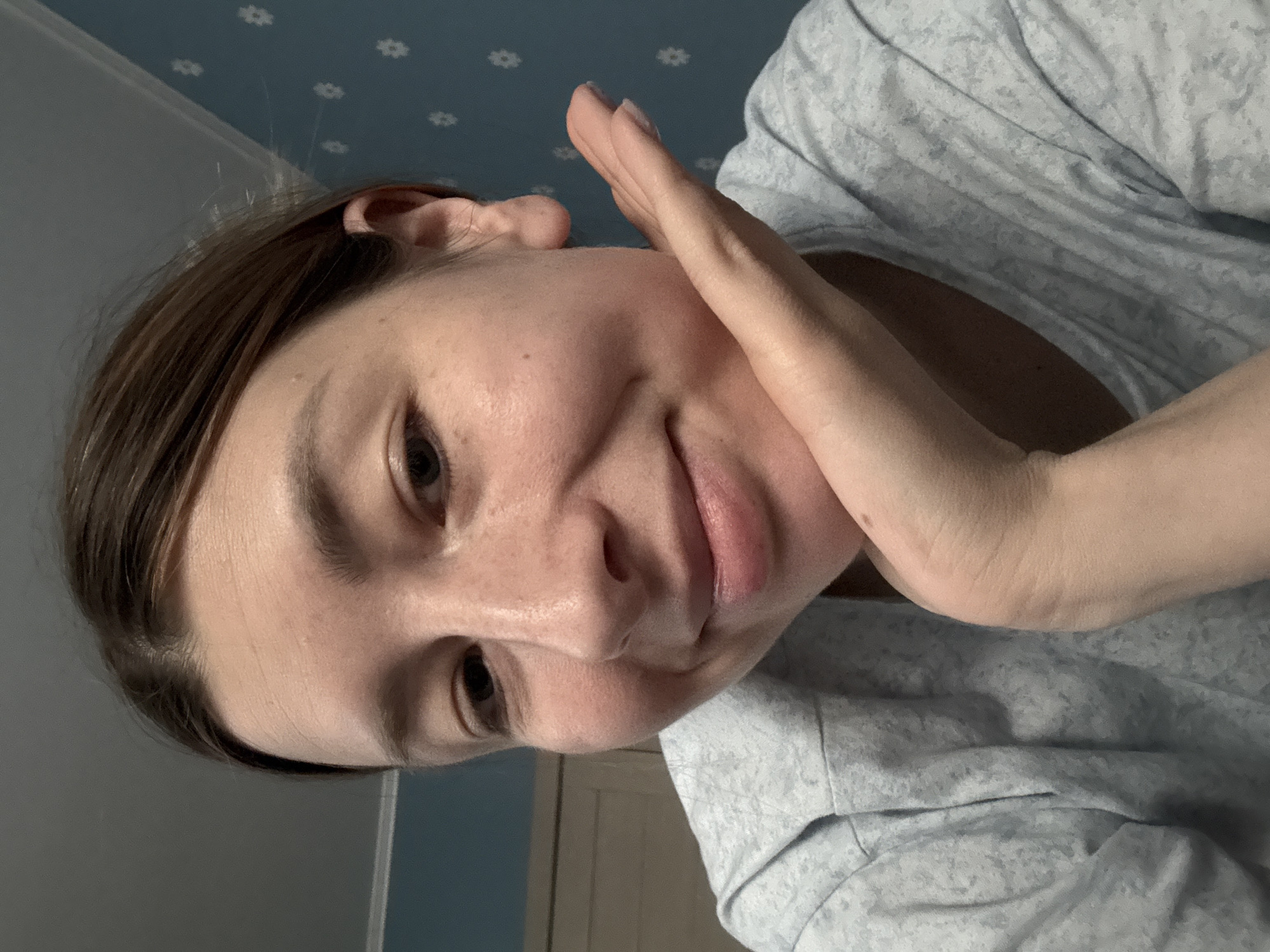 A woman is posing for a selfie, holding her hand up near her face. She is wearing a patterned grey shirt. The background consists of a wall with a floral pattern. There is no evidence to suggest this image was taken in Moscow, Russia.