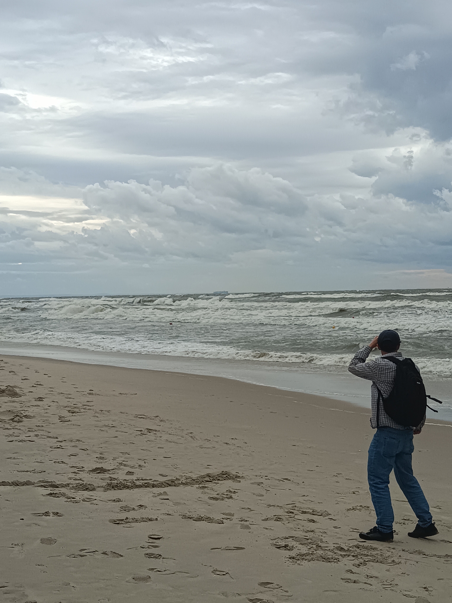 A person stands on a sandy beach facing the choppy sea under a cloudy sky. The individual is wearing a plaid shirt, blue jeans, and a backpack, and is shielding their eyes as if looking out at the horizon. The beach is largely empty, with footprints visible in the sand. The sea is rough with large waves and foam. The sky is overcast with heavy clouds, suggesting a moody or stormy atmosphere. A distant ship is visible on the horizon. The scene is set in Stegna, Poland, a location known for its beaches on the Baltic Sea coast.