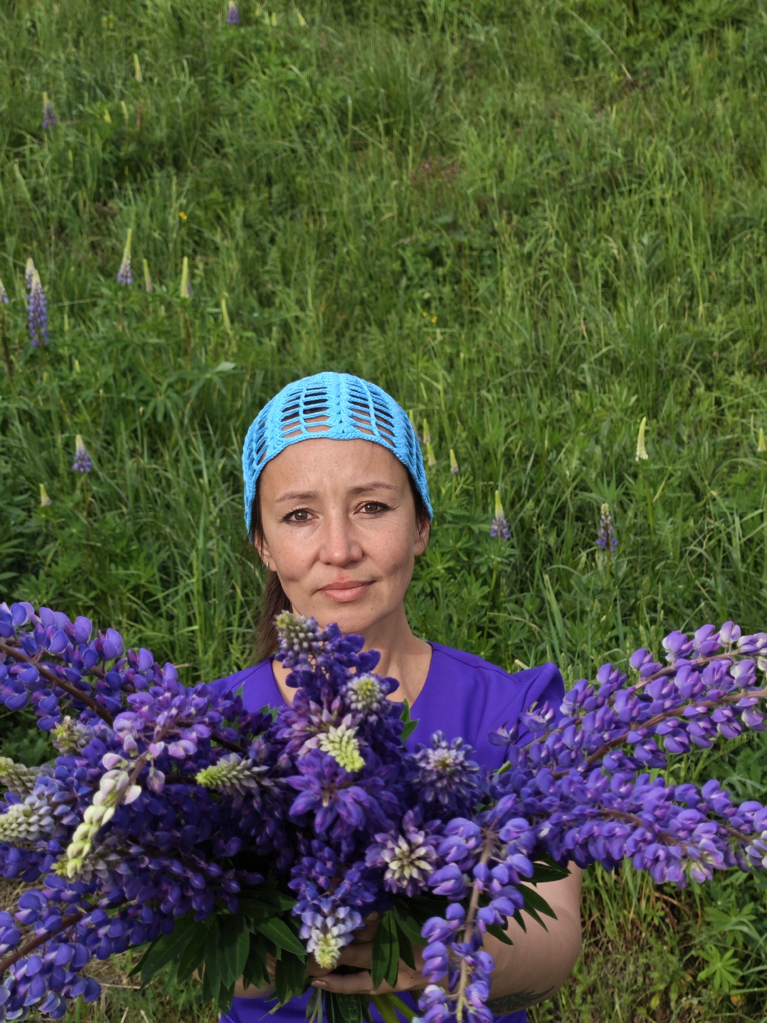 A medium shot depicts a woman holding a large bouquet of purple lupine flowers. She wears a purple shirt and a light blue, crocheted head covering. The woman is centered in the frame, and behind her is tall, green grass with scattered lupine flowers. The photo was taken in the city of Chaikovsky, Russia.