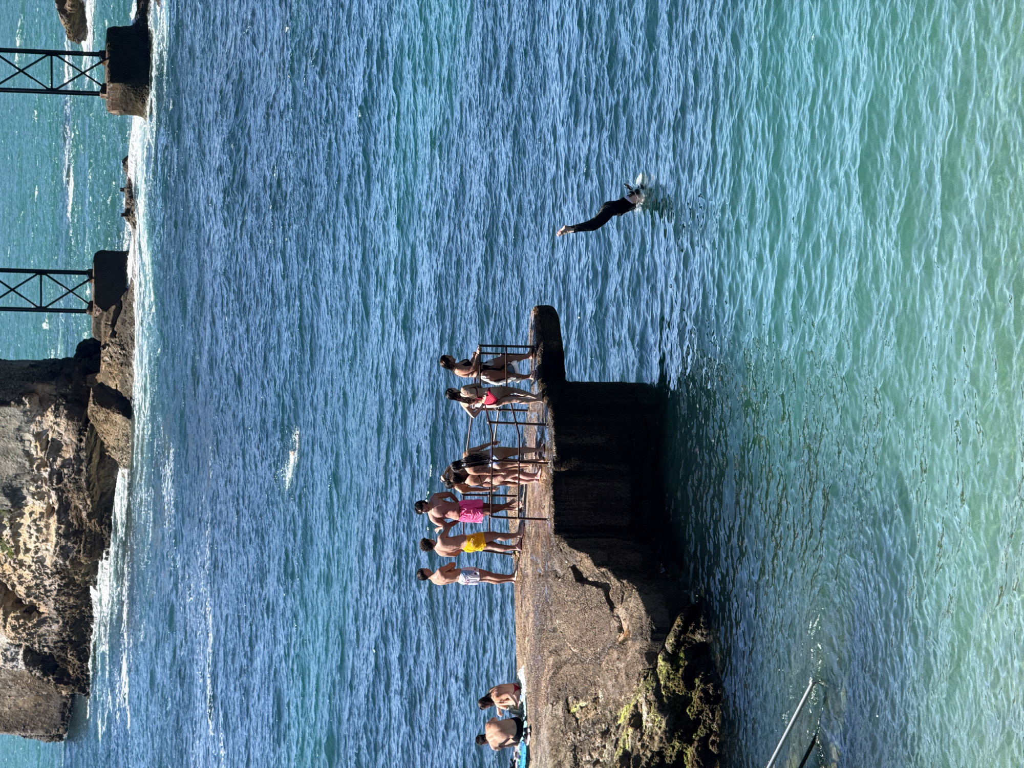 In Biarritz, France, on a sunny day, a group of people are gathered on a concrete pier jutting into the turquoise ocean. Some individuals are standing and conversing on the pier, while others are seated. A person in black swimwear is captured mid-dive into the water from the edge of the pier. The background features large rock formations and industrial-style metal structures in the distance, characteristic of the Biarritz coastline. The water is clear and shows visible ripples.