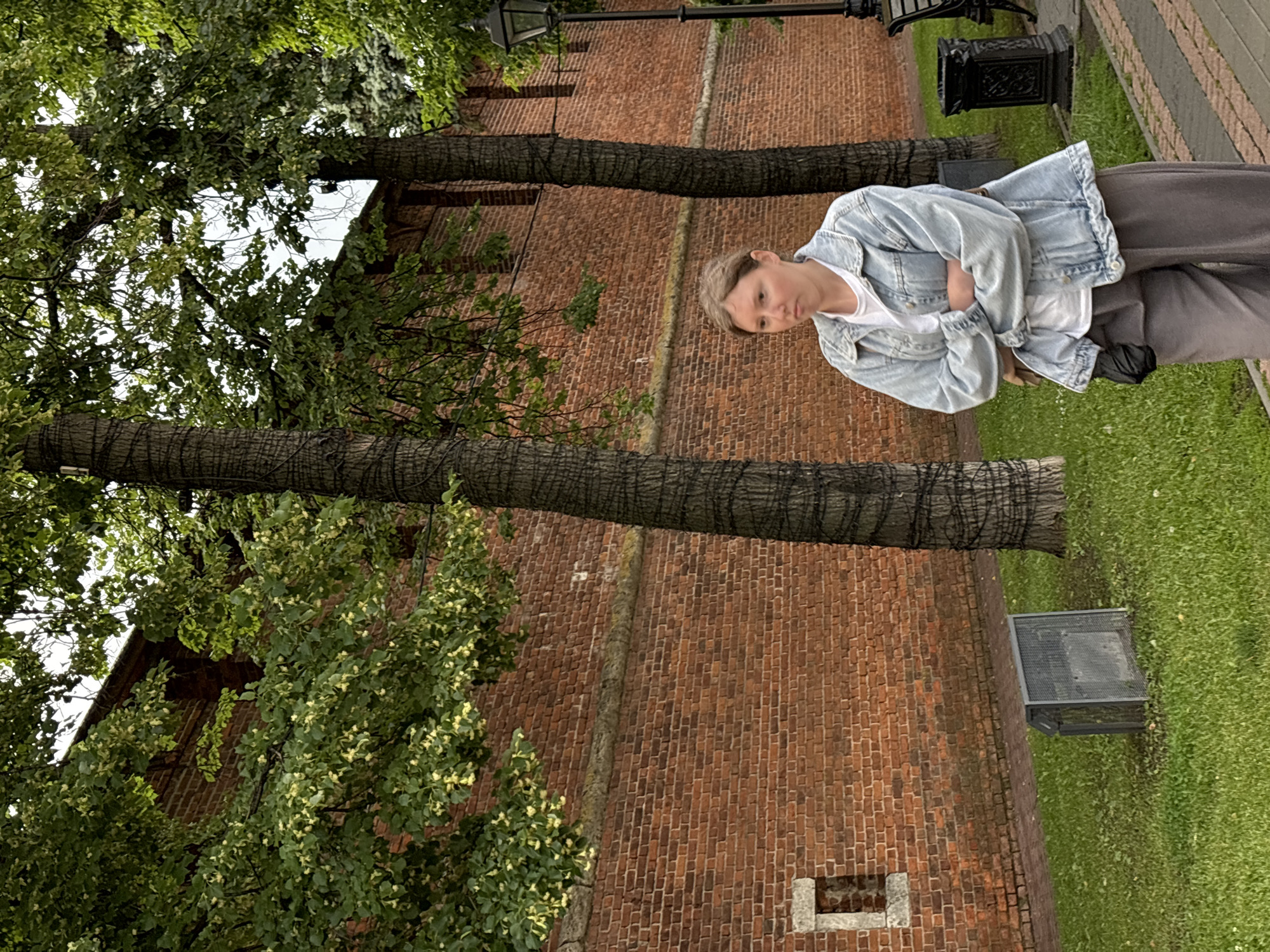A young woman with light brown hair is standing with her arms crossed in front of a brick wall. She is wearing a light blue denim jacket over a white t-shirt and gray pants. The wall is part of the Nizhny Novgorod Kremlin, a historic fortress located in Nizhny Novgorod, Russia. The wall is made of red brick and has a decorative stone band running horizontally across it. Two large trees with dark bark stand in front of the wall, and their branches, covered in green leaves, extend over the scene. The ground in front of the wall is covered in grass, and a cobblestone path runs alongside it. A lamppost and a trash can are visible to the right of the woman. The overall impression is one of a serene, historical setting.
