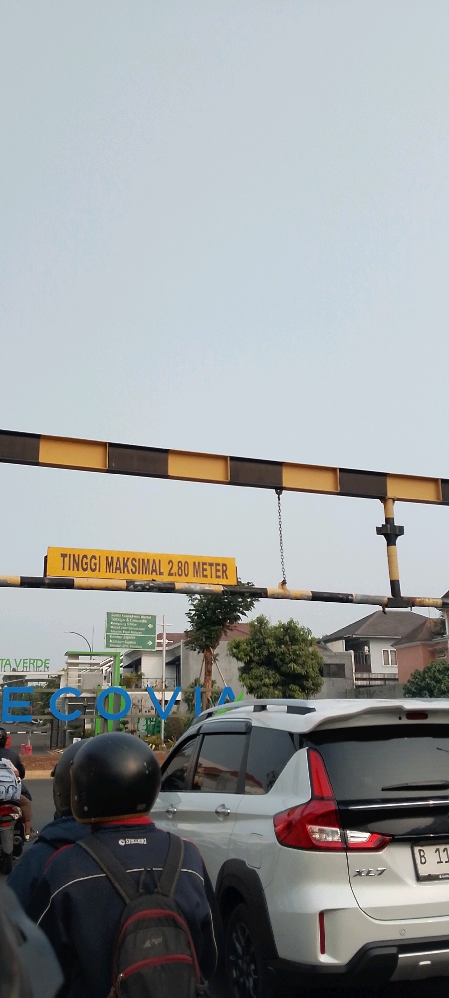 The image captures a street scene in Gunung Putri, Indonesia, with a yellow and black striped overhead barrier. A yellow sign attached to the barrier reads "TINGGI MAKSIMAL 2.80 METER", indicating a maximum height limit of 2.80 meters. Below the barrier, a white SUV with a black spoiler is visible, along with a white license plate starting with "B 11". In the foreground, two individuals on a motorcycle are present, one wearing a black helmet and a backpack. To the left, a large sign with "ECO VIA" written in blue letters can be seen. The environment appears to be an urban or suburban area with buildings and trees in the background, under a bright, overcast sky.