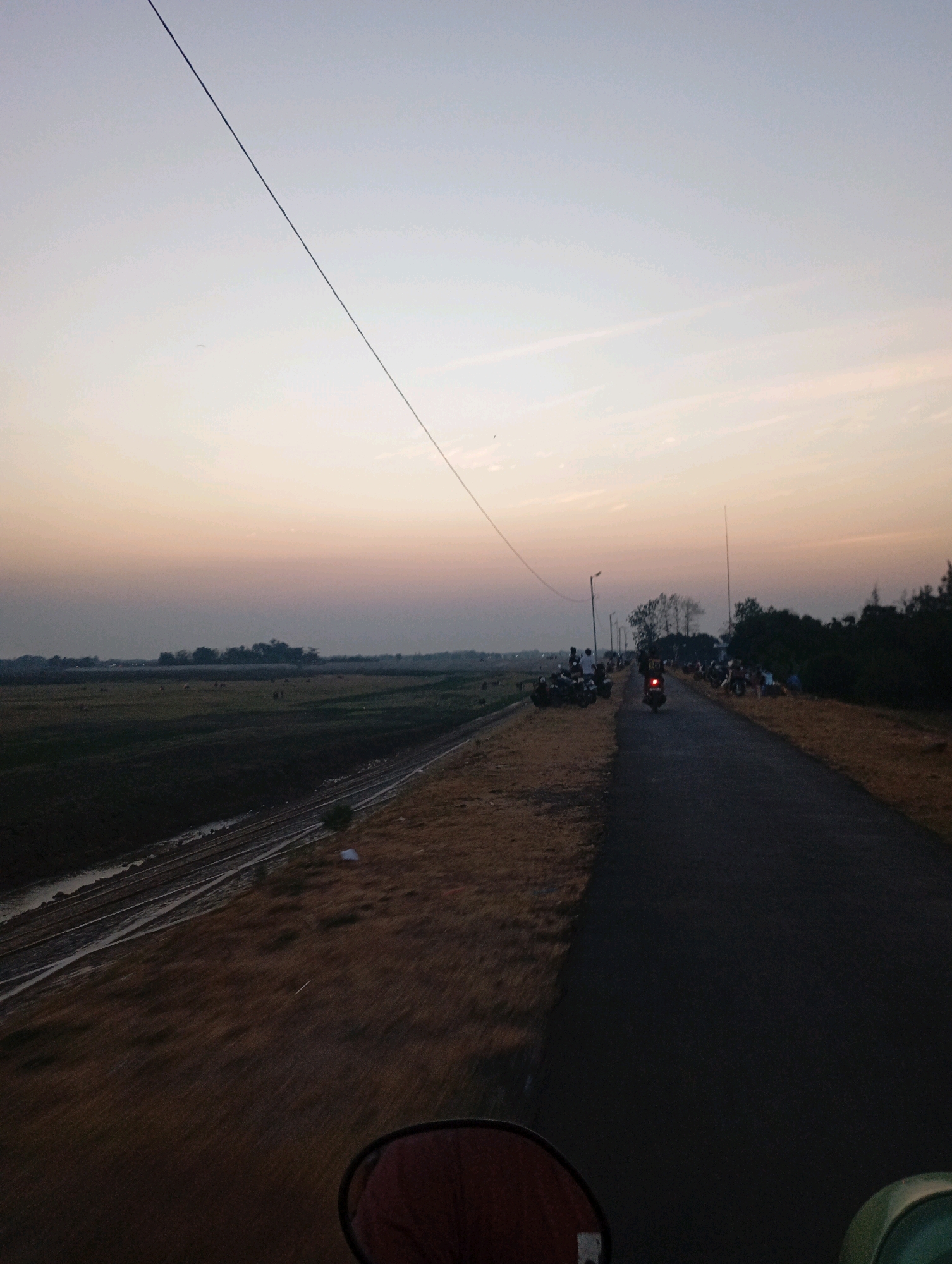 In Haurkolot, Indonesia, a scenic view unfolds at dusk. A paved road stretches into the distance, flanked by dry, grassy terrain on the right and a field of dark green vegetation on the left. A narrow body of water runs parallel to the field. The sky displays a soft gradient of pale blue and peach, with a thin, dark power line cutting diagonally across it. Along the roadside, several people are gathered, some standing near motorcycles, while others are seated. One individual is riding a scooter away from the viewer, its brake lights illuminated. In the background, a line of trees marks the horizon, and a cluster of hay bales is visible in the field. The scene is captured from a low vantage point, possibly from a vehicle, with the reflection of a red object in a round mirror visible at the bottom of the frame.