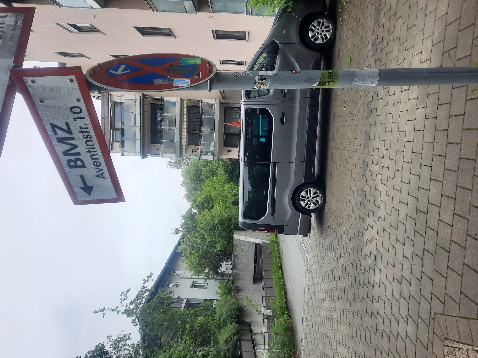 A gray van is parked on a cobblestone street in front of a light pink building. A street sign indicates "BMZ Aventinstr. 10" with an arrow pointing left. A circular traffic sign with a red border and blue and red markings is attached to a pole. Trees and foliage are visible on the left side of the image, and the sky is overcast.