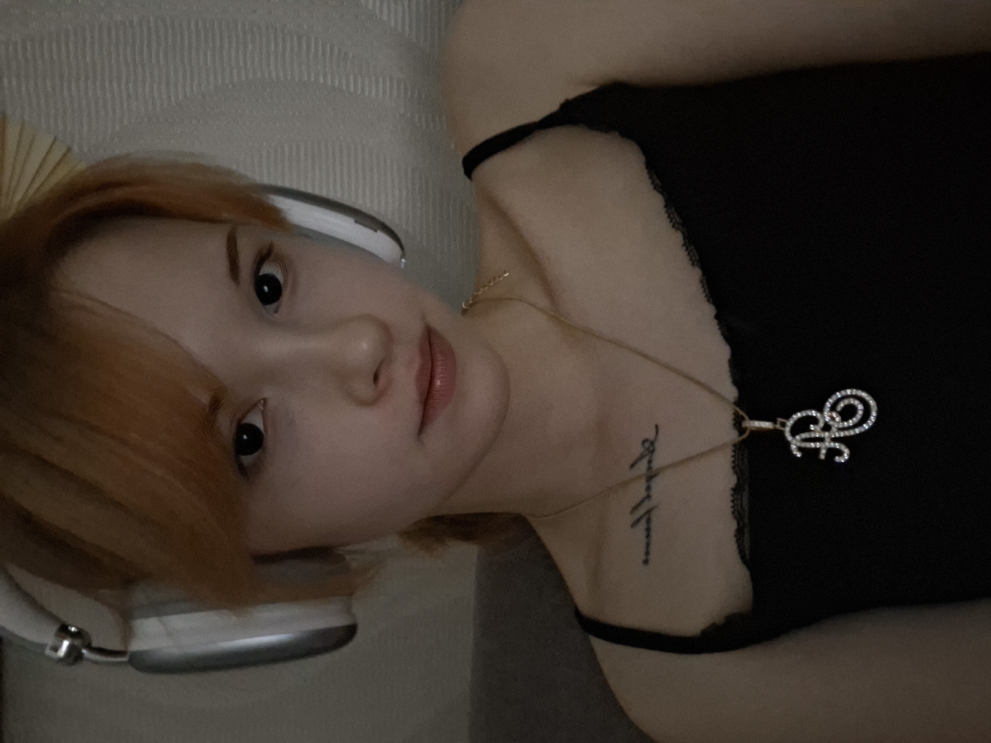 A young woman is captured in a portrait-style photograph, lying down indoors and looking directly at the camera. She has shoulder-length, reddish-orange hair that frames her face, and fair skin with dark, attentive eyes. Her expression is calm and neutral. She is wearing a black camisole or tank top featuring delicate lace trim along the neckline.
Over her ears, she wears white over-ear headphones, resembling Apple AirPods Max, suggesting she is engaged in listening to music or audio. Adorning her neck is a thin gold chain necklace from which hangs a sparkling, ornate pendant in the shape of a stylized letter 'F', possibly encrusted with small gemstones. On her right collarbone, a script tattoo is visible, appearing to be two words that are difficult to fully decipher but resemble a phrase like 'Always Forever'.
The background is a simple, light-colored textured surface with vertical patterns, possibly a headboard or a wall. The lighting is soft and ambient, suggesting an indoor setting, likely in the evening. The overall scene conveys a moment of relaxed leisure in Chaikovsky, Russia.