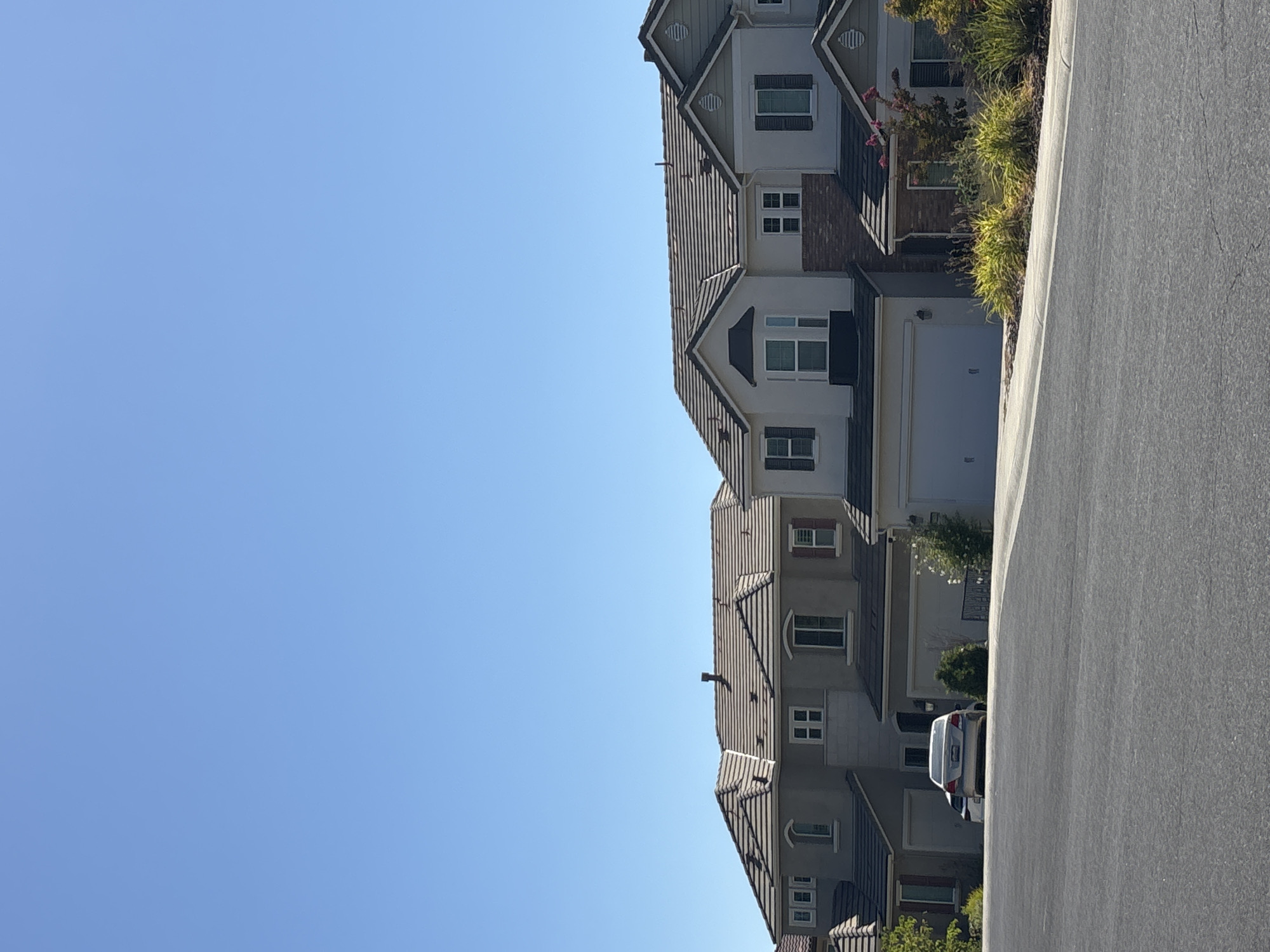 In the city of Roseville, California, United States of America, a suburban street scene unfolds under a clear blue sky. The foreground features an asphalt road with a white painted line on the right side, curving gently towards the middle ground. On the left side of the road, a light-colored sedan is parked. Beyond the road, a row of attached houses, each with a two-car garage, stretches across the frame. The houses are two stories tall and feature beige stucco exteriors with dark tiled roofs. Some houses have dark shutters on their windows and red brick accents around the garages and entrances. Lush green bushes and some flowering plants line the front of the houses, adding a touch of nature to the urban landscape. The scene is peaceful and serene, typical of a residential neighborhood.