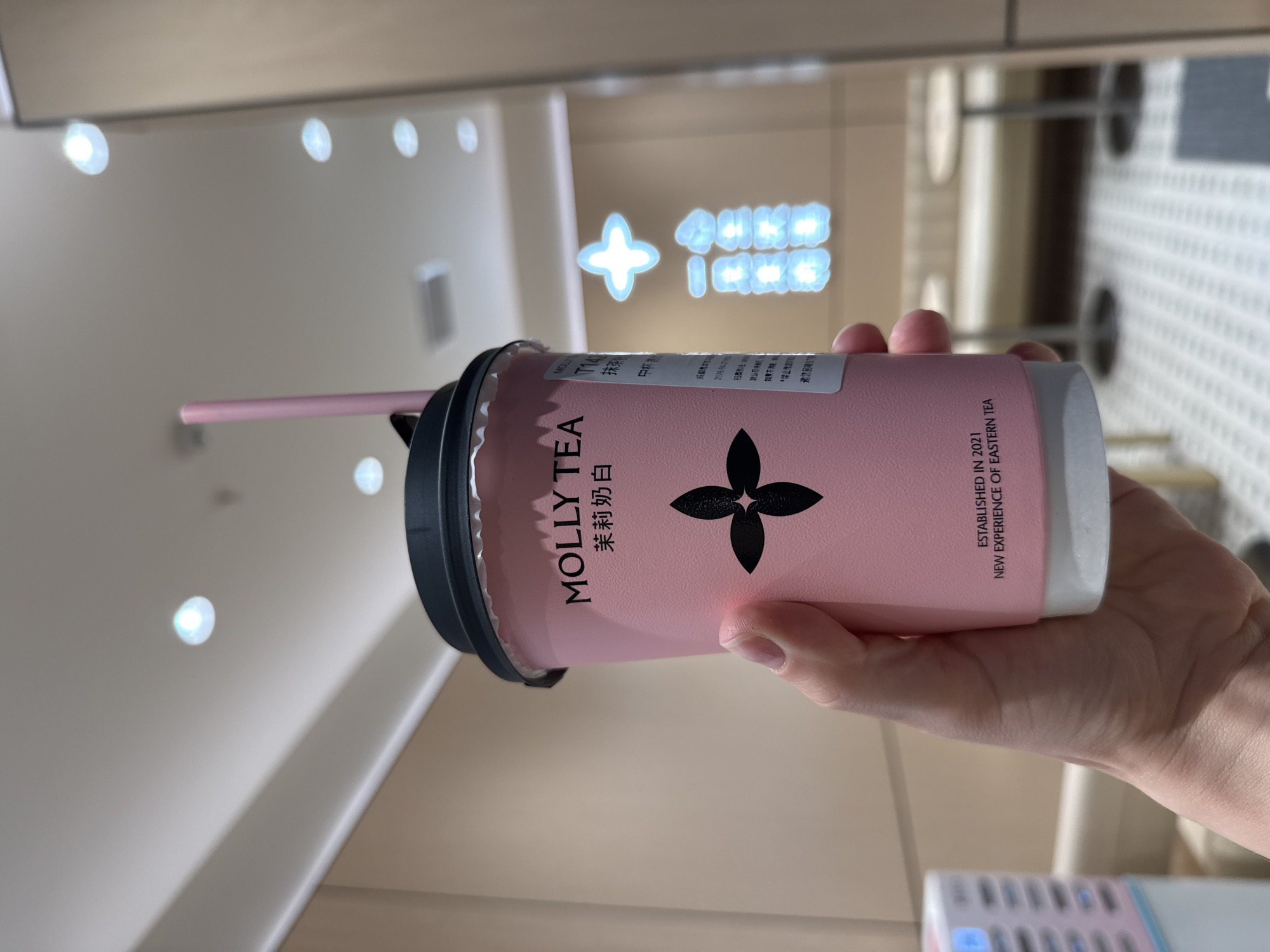 A hand holds a pink beverage cup with a black lid and a pink straw, captured from a slightly elevated angle. The cup is designed with a prominent black floral or clover-like logo and text. Visible branding on the cup includes "MOLLY TEA," "茉莉奶白" (Jasmine Milk White or Jasmine Milk Tea), "ESTABLISHED IN 2021," and "NEW EXPERIENCE OF EASTERN TEA." Other smaller Chinese characters like "抹茶" (Matcha), "20960417", "如果", "禁止", and "邀您品味" (Invite you to taste/savor) are also partially discernible.
The scene is indoors, likely a public area such as a commercial building or shopping mall in Qingyang District, China. The background features a light-colored ceiling with multiple bright, circular overhead lights, creating bokeh effects. In the mid-ground, a luminous sign with a glowing cross-like symbol and blurry Chinese characters is visible. The image conveys a clean, modern aesthetic. The time of day is not explicitly indicated but appears to be well-lit indoors. The focus is primarily on the branded beverage cup.