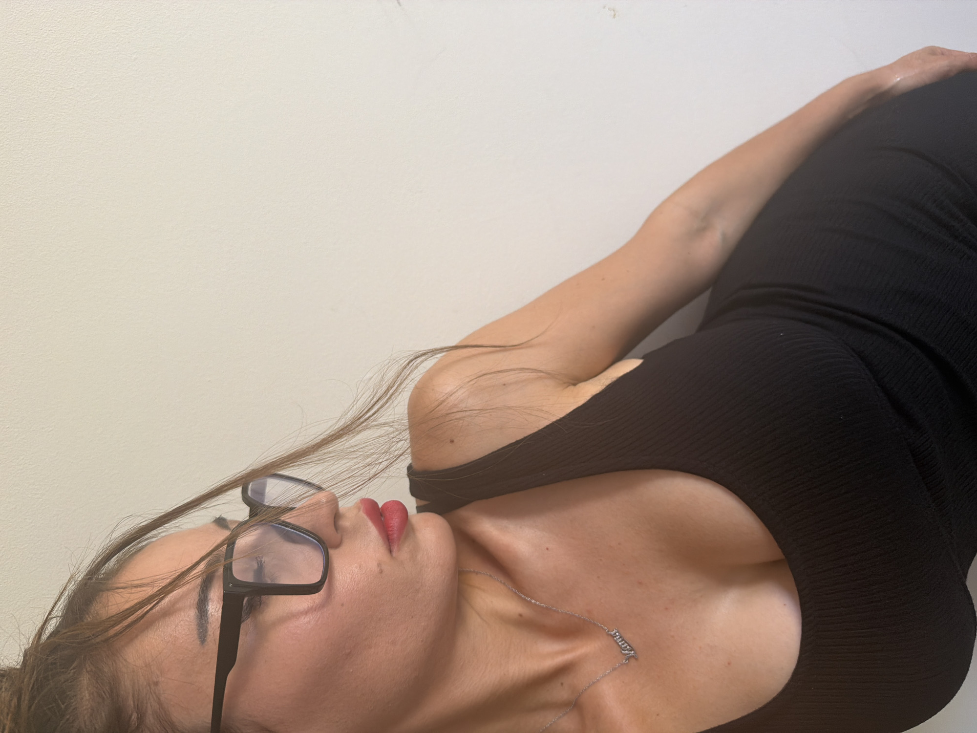A medium shot shows a woman with brown hair pulled up, wearing black-rimmed glasses and a black sleeveless dress. She is looking to her left, with her right arm resting on her right thigh. A silver necklace with a pendant spelling "Mama" is visible on her neck. The background is a plain, off-white wall, consistent with an indoor setting. The image appears to be taken in Tersefanou, Cyprus.