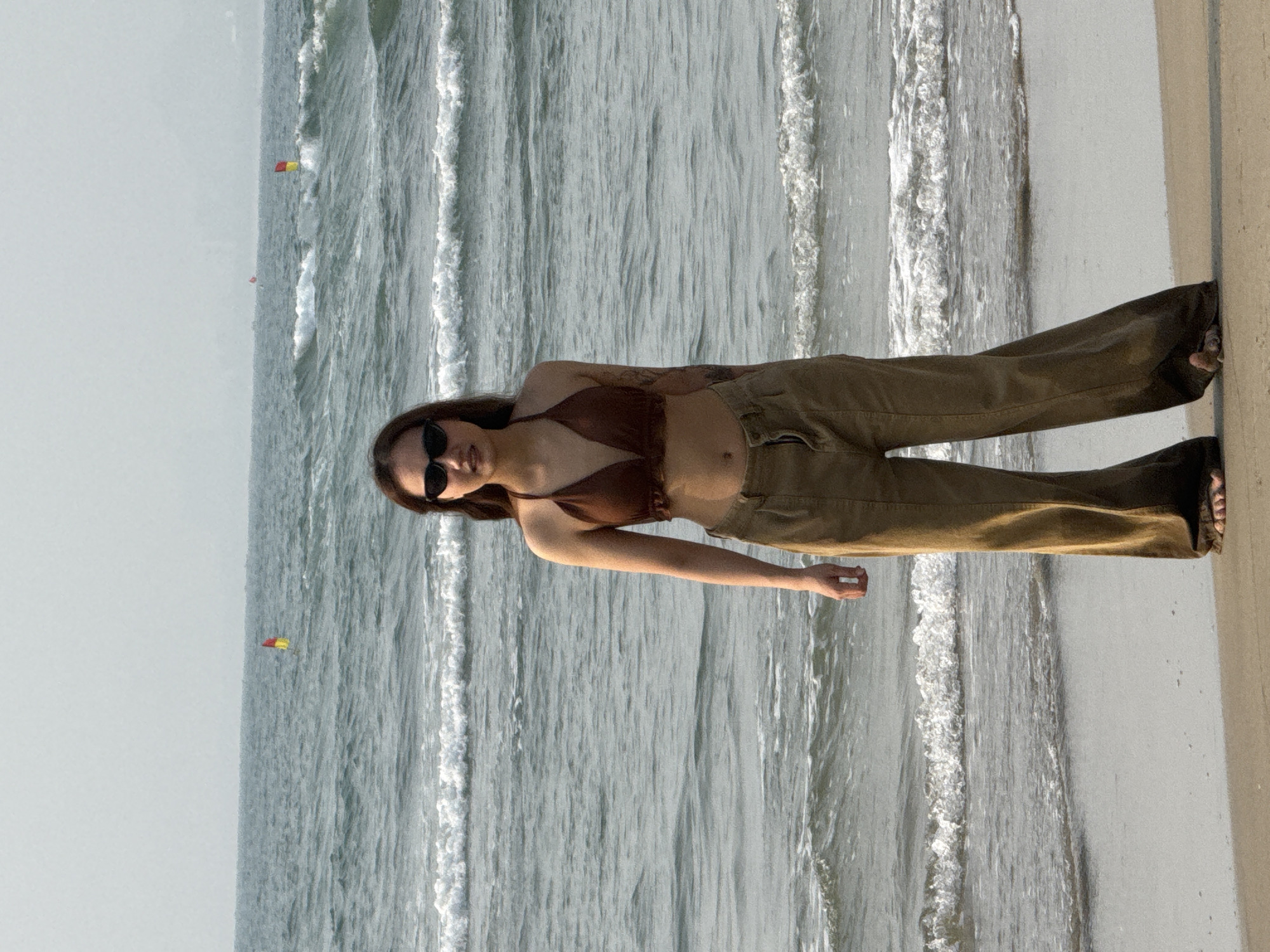 A young woman wearing a brown bikini top, brown pants, and sunglasses stands on a sandy beach next to a body of water with small waves. Kites are flying in the distance. The location is Đà Nẵng, Vietnam.