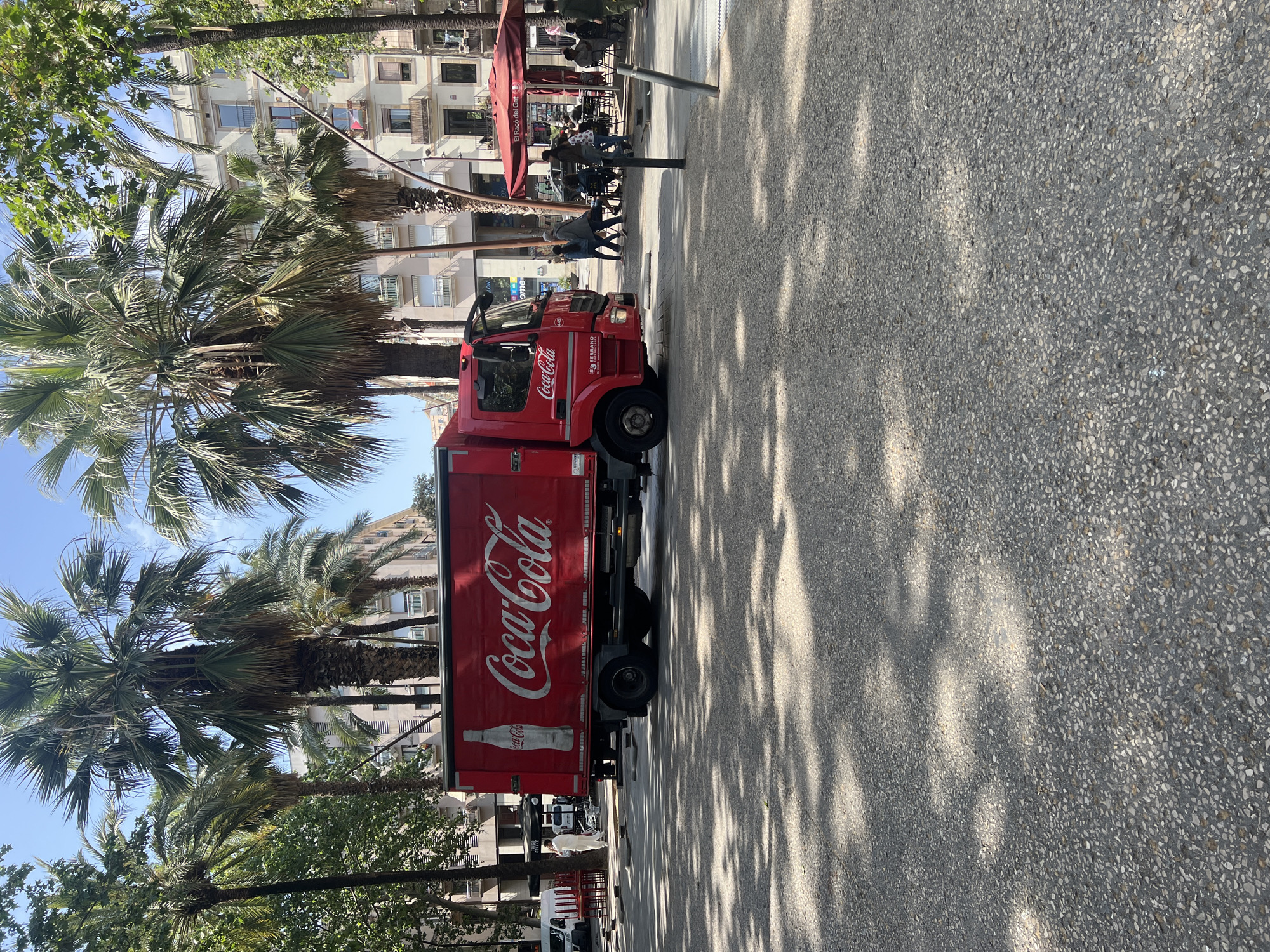 This image captures a vibrant urban street scene in Barcelona, Spain, under bright daylight. A large red Coca-Cola delivery truck is prominently parked on the right side of a wide, paved street or pedestrian area. The truck features the classic "Coca-Cola" logo in white script along its entire side, and smaller details like "Coca-Cola," "SERRANO," and the number "445" are visible on its cab.
The left side of the street is lined with several tall palm trees and other green trees, which cast distinct, dappled shadows across the light grey, speckled pavement. In the background, multi-story buildings with balconies and numerous windows complete the urban landscape. To the far left, an outdoor cafe or restaurant area is visible, distinguished by red awnings and umbrellas under which several people are seated at tables. The name "El Racó del Gat" (Catalan for "The Cat's Corner") and partially "COFFEE" are legible on one of the red awnings.
The scene depicts typical daily activities: people are walking on the pavement, while others are relaxing and socializing at the outdoor cafe. The bright sunlight and clear blue sky suggest a pleasant, warm day, likely late morning or early afternoon. The image offers a glimpse into the everyday rhythm of a European city, combining commercial activity with leisure in a lively setting.
