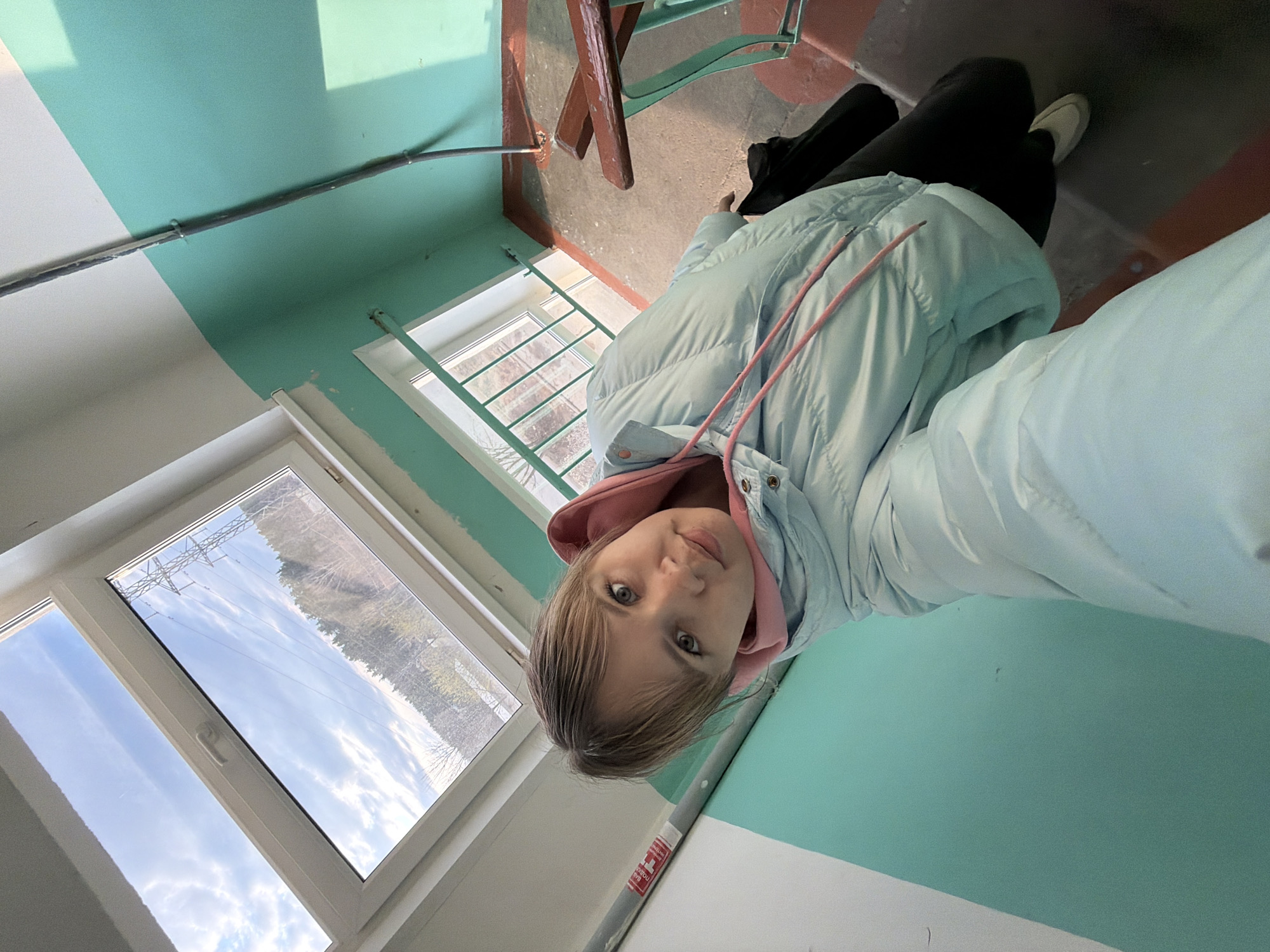 A young person is captured in a high-angle selfie or photo, appearing to be seated or crouching on the floor of what resembles a utilitarian indoor space, possibly a stairwell or hallway. The person, with light brown hair and light eyes, looks directly at the camera with a neutral expression. They are wearing a light blue puffer jacket over a pink hooded sweatshirt, black pants, and light-colored sneakers.
The interior features walls painted in a distinct two-tone style: a lower section of white and an upper section of light greenish-blue (aqua or teal). A dark pipe or conduit runs along the wall where the two colors meet. The floor is a rough, concrete-like surface, with a reddish-brown painted trim visible. A wooden handrail, possibly part of a staircase, is also seen.
To the left, a large window allows ample daylight to stream in, illuminating the scene. Through the window, an outdoor environment is visible, featuring trees and power lines or towers against a partly cloudy sky. The overall impression is one of a bright, clear day.
On a grey pipe in the foreground, there is a white rectangular sticker with red text. While partially obscured, the text appears to be in Cyrillic, with "НЕ" clearly visible, suggesting a warning or instruction, possibly "НЕ ПОДКЛЮЧАТЬ!" (DO NOT CONNECT!).