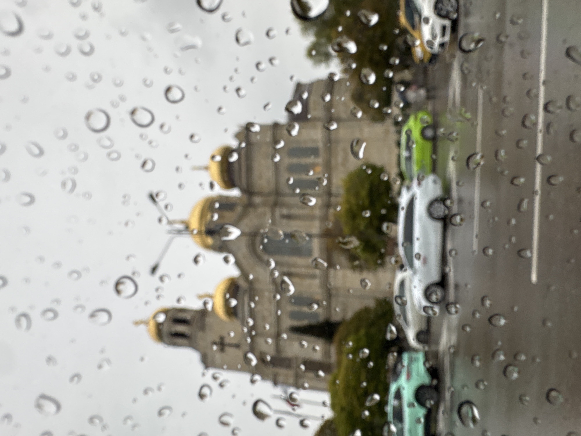 The image presents an outdoor scene viewed through a surface heavily covered with water droplets, indicating rain or recent precipitation. These droplets are in sharp focus in the foreground, creating a significant bokeh effect that blurs the background elements.
In the mid-ground and background, a majestic building with a grey stone facade and multiple prominent golden domes is visible, occupying the left side of the frame. Its architectural style suggests a large cathedral or church, consistent with structures like the Dormition of the Theotokos Cathedral in Varna, Bulgaria, which aligns with the provided location context. The building appears ornate, featuring arches and intricate details, though these are softened by the blur and water droplets.
To the right of the building, a street can be seen with several cars parked alongside it. A vibrant green car is clearly distinguishable, followed by a lighter-colored vehicle, possibly white or silver. Another car, partially visible at the top, has yellow and white elements. Green trees are interspersed between the building and the street, adding hints of nature to the urban environment.
The time of day appears to be daytime, under overcast or rainy conditions, contributing to the diffused and somewhat muted lighting of the scene. The image captures a static, contemplative moment, observed from behind a rain-splashed window, showcasing an urban landscape in Varna, Bulgaria, during inclement weather. There is no visible text or human activity depicted.