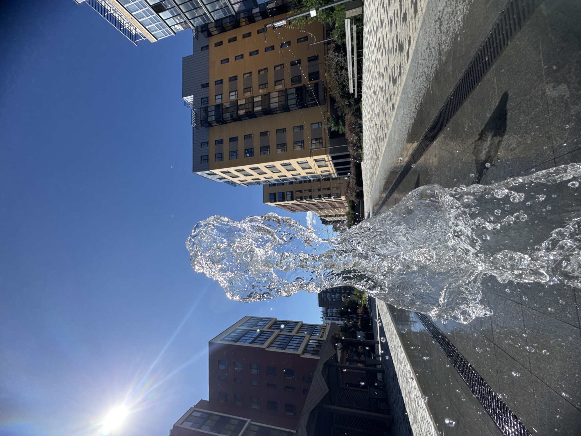 The image captures a close-up view of a water fountain in the city of Novoselye, Russia. The fountain's water spray is in the foreground, frozen in mid-air and catching the sunlight. Behind the fountain, a modern urban landscape unfolds with several multi-story buildings. The buildings are constructed with brick and glass facades, featuring balconies and a clean, contemporary architectural style. A paved pedestrian area with intricate tiling runs in front of the buildings. The sky above is a clear, vibrant blue, with the sun brightly shining and casting lens flares into the shot. The scene is set on a sunny day, emphasizing the refreshing aspect of the water feature against the urban backdrop.