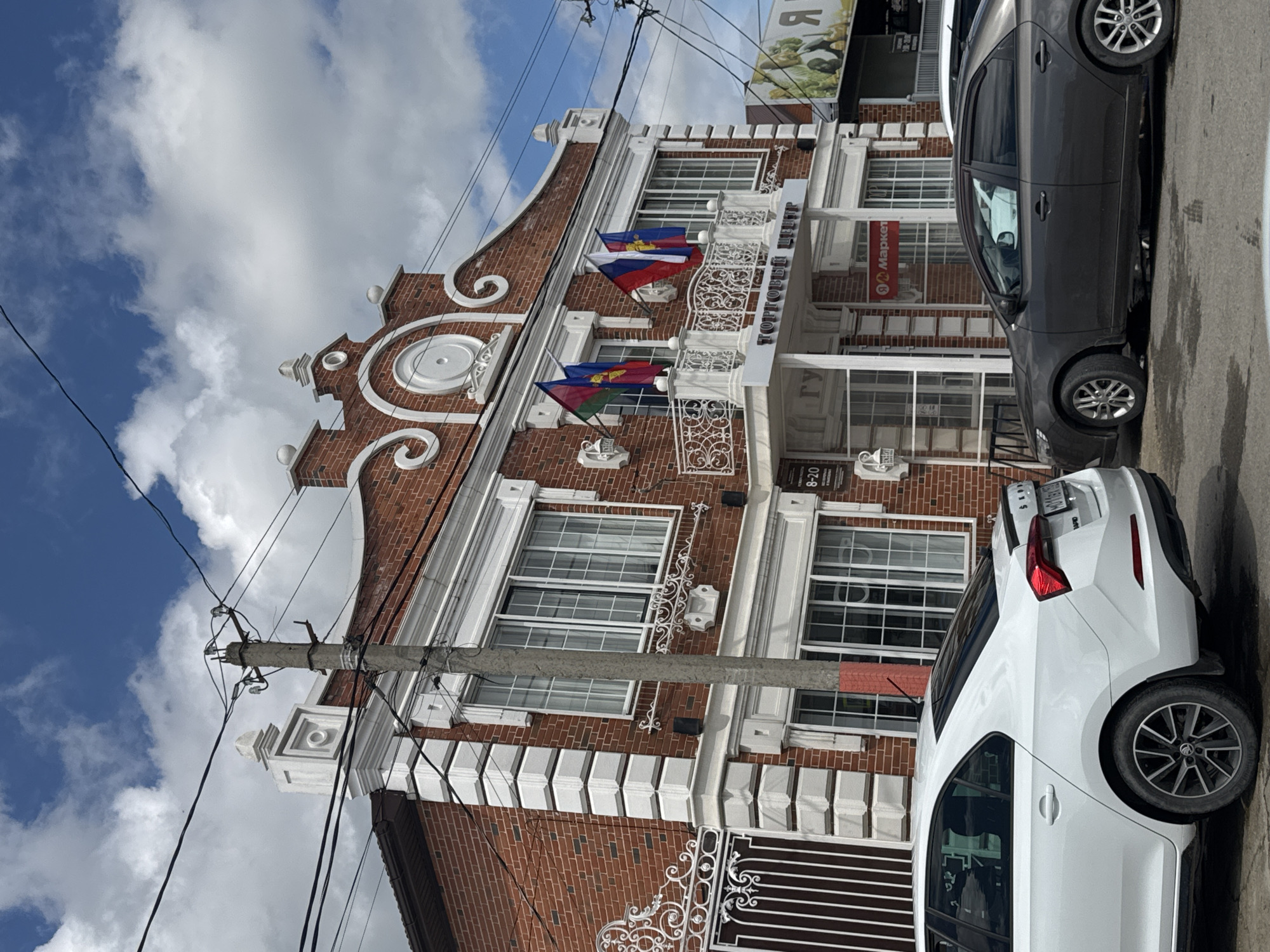 The image shows a three-story brick building in Yelizavetinskaya, Russia, with distinctive Dutch gable architecture. The building has white trim and decorative wrought iron elements. Two cars are parked in front of the building: a white MG sedan and a dark gray sedan. The building flies the flags of Adygea. Power lines and a utility pole are visible in the foreground. The sky is partly cloudy.