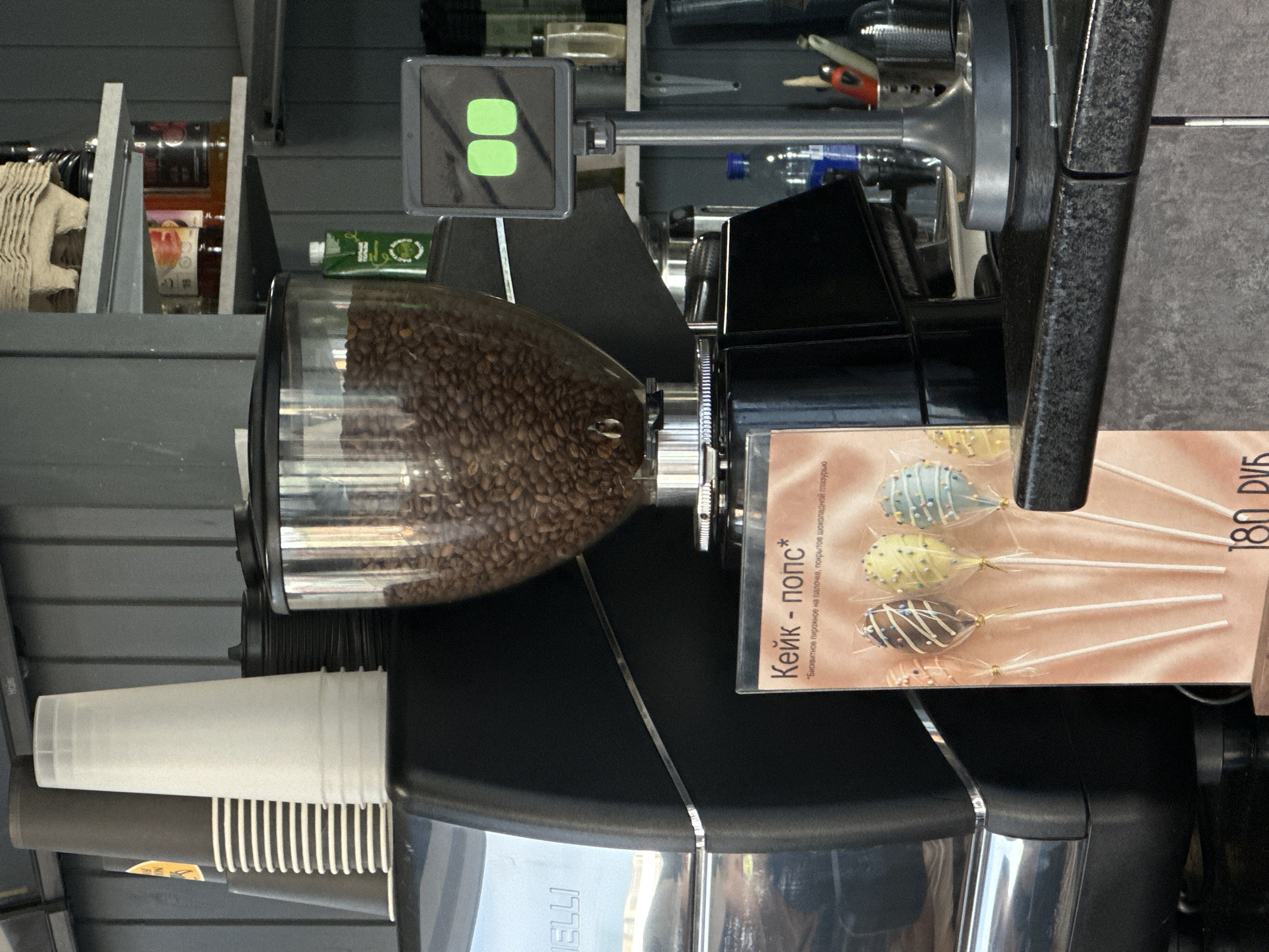 The image captures a close-up view of a coffee station, likely in a cafe or snack bar. The central focus is a professional-grade coffee machine, prominently featuring a clear hopper filled with numerous dark coffee beans, ready for grinding. Below the hopper, part of the machine's metallic base is visible with the partial brand name "ELLI" inscribed.
To the left of the coffee machine, a stack of translucent white plastic cups and another stack of black and white striped paper cups are neatly arranged, awaiting use. On the right, a vertical menu or advertisement card is displayed, showcasing four colorful cake pops on sticks, individually wrapped in clear plastic. These cake pops are decorated with various glazes and sprinkles: one is light blue, another yellow, a third dark brown, and the bottom one appears light pink or peach.
Visible text on the card reads: "Кейк - попс*" (Cake - pops*) followed by a descriptive asterisked phrase "*Бисквитное пирожное на палочке, покрытое шоколадной глазурью" (*Biscuit cake on a stick, covered with chocolate glaze). The price "180 РУБ" (180 RUB, likely Russian Rubles) is also clearly shown.
Above the coffee machine, a square tablet-like display is mounted, showing two prominent bright green square icons on a dark screen. In the background, shelves are visible, holding various bottles and containers, including one green-packaged item. The overall setting appears clean and functional, with dark surfaces and metallic accents, suggesting an indoor commercial environment ready for serving customers.