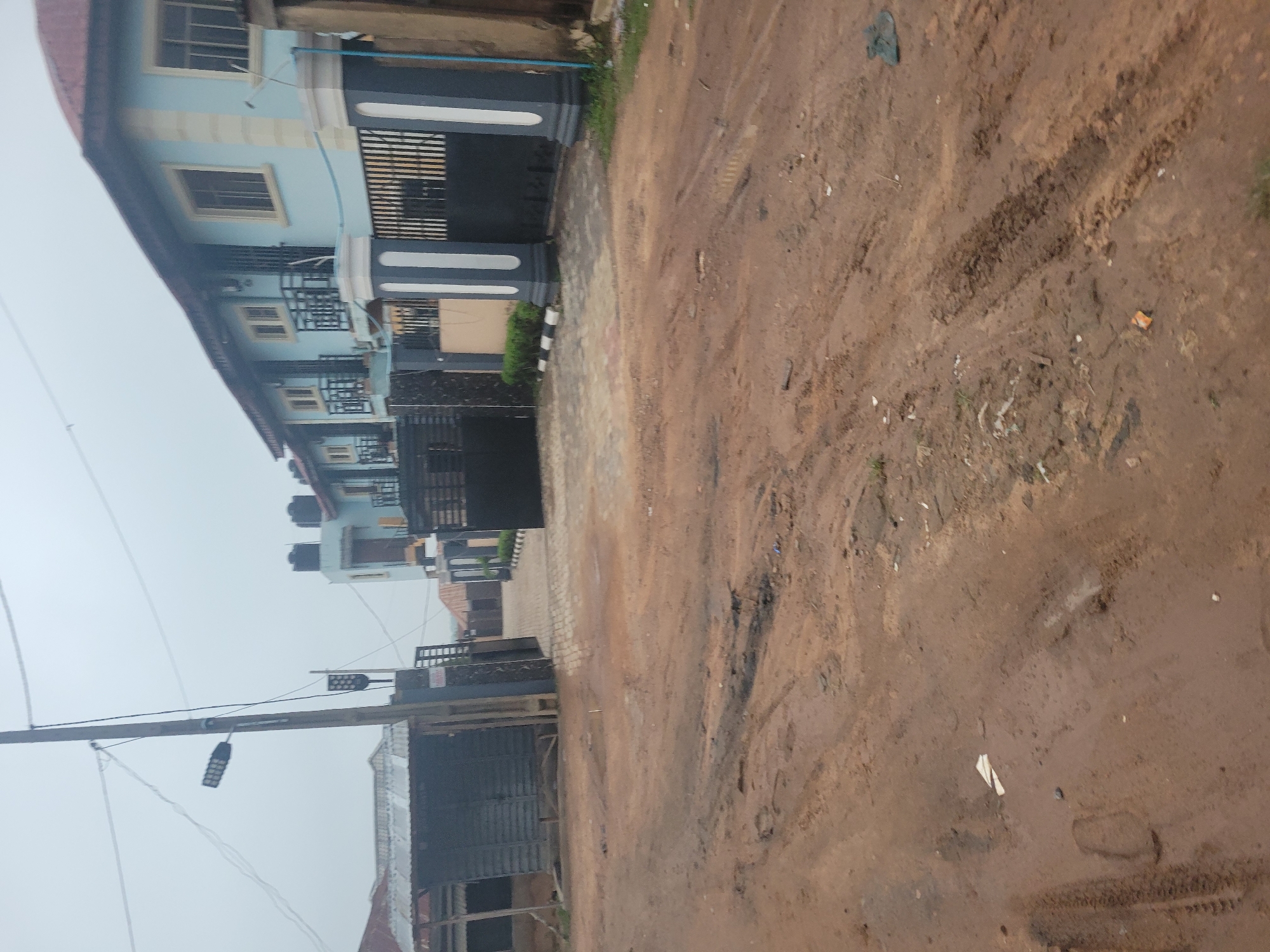 The image shows a street in a city. The street is muddy and appears to be unpaved, with tire tracks and footprints visible. There are several buildings in the background, including a light blue multi-story building with a red roof, and a smaller dark-colored structure with a metal roof. A large black metal gate and decorative pillars are also visible. A utility pole with two lights is on the left side of the image. The sky is overcast.