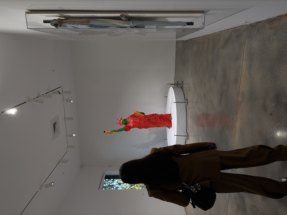 The image depicts an interior view of a modern art gallery, identified as the Rubell Museum in Miami, Florida. The room features white walls, a high ceiling with exposed track lighting and an air vent, and a grey concrete floor. A tall window on the left wall shows green foliage and blue sky outside. A security camera is mounted high on the left wall.
In the foreground, slightly to the left, a person is seen from behind, facing away from the viewer towards the exhibits. This individual has long, dark hair and is wearing a dark brown two-piece suit consisting of a jacket and wide-legged trousers. They carry a black shoulder bag on their left side, and a blue-cased smartphone is partially visible in their right hand. White athletic shoes are visible on their feet.
The central subject of the room is a large, colorful sculpture of the Statue of Liberty, positioned on a circular white display pedestal in the middle ground. The statue is predominantly red, covered in a textured or mosaic pattern. Its head, crown, and raised right arm holding the torch are bright green. The torch contains a yellow-orange flame with a black tip. The pedestal is demarcated by a low barrier of thin metal stanchions.
To the right, mounted vertically on the white wall, is another artwork enclosed within a clear acrylic display case. This piece appears to be a mixed-media composition, featuring light blue fabric resembling denim, along with light brown rope elements, arranged in an elongated form that suggests a deconstructed or folded umbrella. The individual in the foreground is oriented towards these displayed artworks, observing the scene.