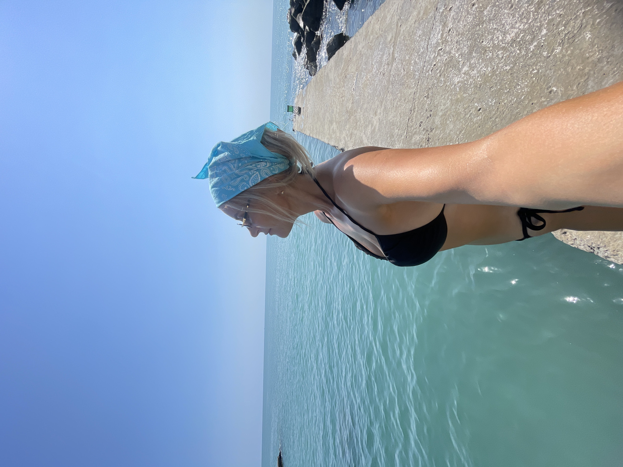 A woman in a black bikini and a blue bandana stands on a concrete pier, looking out at the Black Sea. The water is a turquoise color, and the sky is clear and blue. In the background, there are rocks and the distant outline of the city of Sochi. The image appears to have been taken on a sunny day.