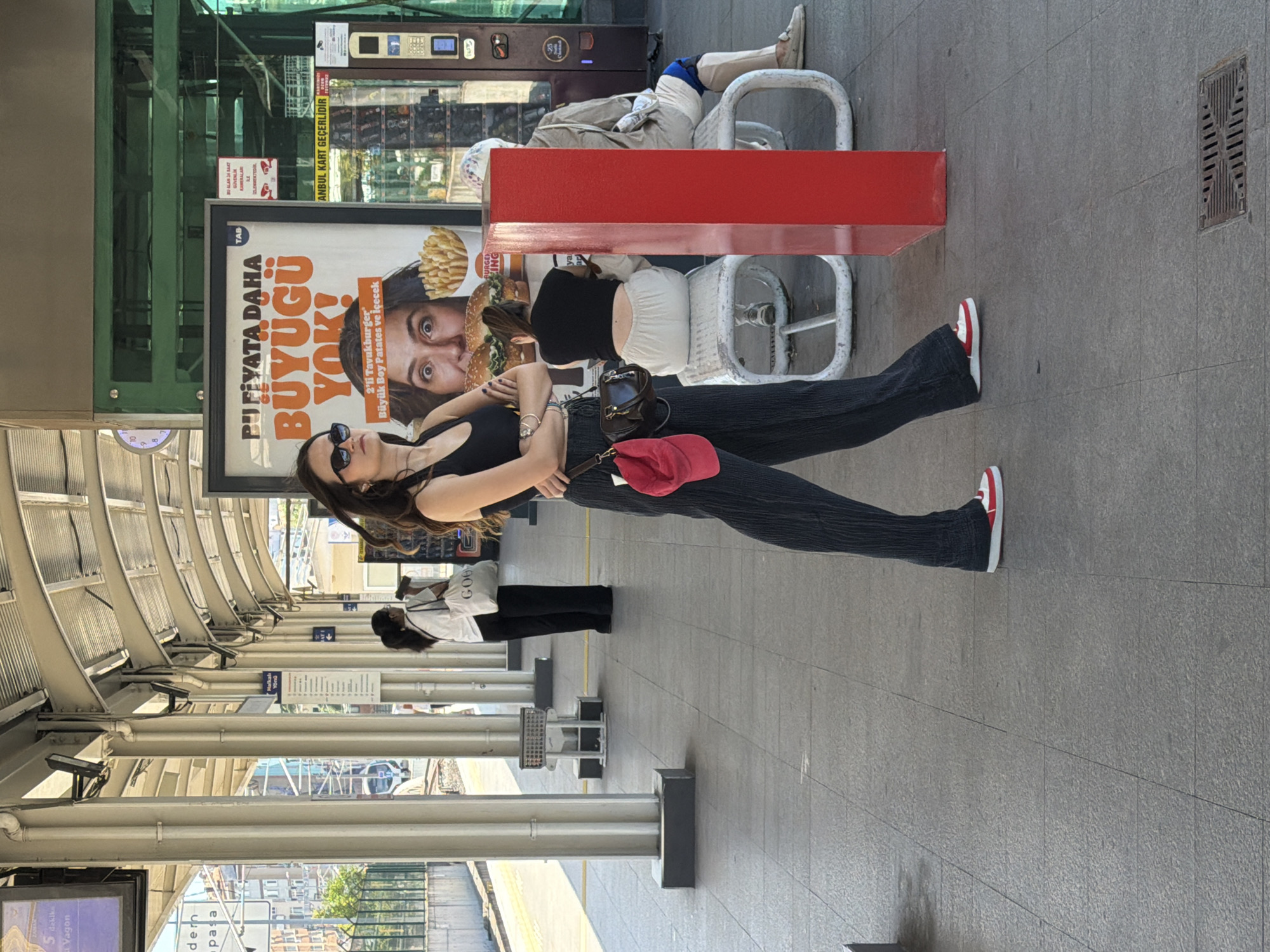 The image captures a sunny outdoor scene at a public transportation station, likely a train or tram stop, in Kadıköy, Turkey. In the foreground, a young woman with dark hair and sunglasses stands with her arms crossed, wearing a black crop top, dark flared pants, and red and white sneakers. A red cap is slung over her shoulder, attached to a brown crossbody bag. Behind her, another woman in black pants and a white t-shirt carries a white tote bag. Further back, two individuals are seated on a white bench. One is a person wearing a light brown jacket and light-colored pants, and the other is a person in a black top and light-colored pants. A large advertisement for Burger King is prominently displayed, featuring a close-up of a woman's face and images of burgers and fries. The station has a covered walkway supported by metal pillars, and the ground is paved with grey tiles. The overall atmosphere suggests a typical day at a bustling urban transit hub.