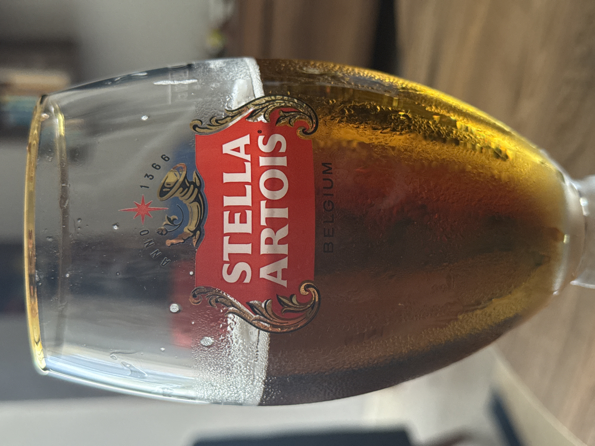 A close-up, rotated shot focuses on a glass of Stella Artois beer. The glass is mostly empty, with a small amount of golden-brown beer remaining at the bottom. Condensation is visible on the outside of the glass, indicating it is cold.
A prominent red label with white lettering reads "STELLA ARTOIS" vertically. Above and below the text are ornate golden flourishes. To the left of the main label, a circular emblem features a starburst, the number "1366", and a trumpet or horn-like symbol. The word "ANNO" is visible in an arc above the emblem, and a faint "BELGIUM" is printed vertically to the right of the "STELLA ARTOIS" text.
The background is blurred, showing warm-toned wooden surfaces and suggesting an indoor setting. The lighting appears to be natural, possibly daylight, casting a warm glow on the beer. No people are visible in the frame, and the overall mood is casual and relaxed, centered around the enjoyment of the beverage. The setting is identified as Eyüpsultan, Turkey.