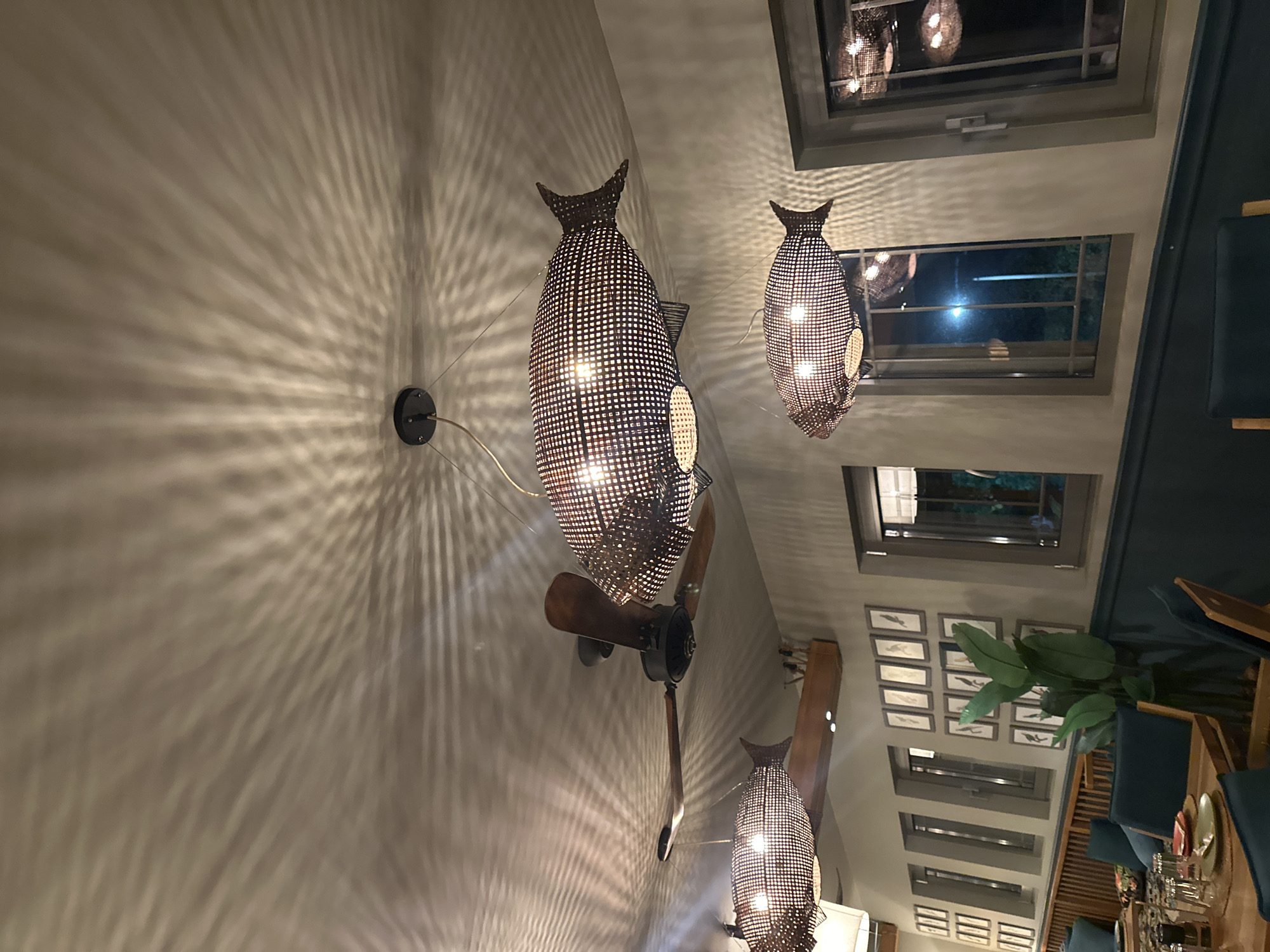 A low-angle shot captures the interior of a dimly lit restaurant with a ceiling fan and decorative fish-shaped lamps. The lamps, made of a woven material with a grid pattern, cast intricate light and shadow designs on the ceiling. A ceiling fan with wooden blades is positioned between two of the lamps. Below, the restaurant features framed artwork on a light-colored wall, a dark green wainscoting, and a wooden bench with teal cushions. Several tables are visible with place settings, including glasses and plates. Windows with grey frames are present on the wall, reflecting the interior lighting and the dark exterior.