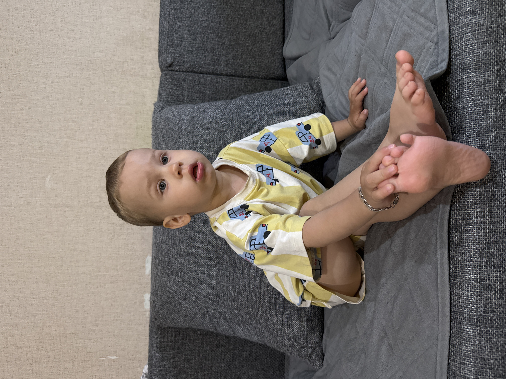 A baby or toddler is pictured sitting or reclining on a grey sofa, looking directly towards the camera with a curious expression and slightly open mouth. The image is oriented sideways, with the baby's head to the right and feet to the left of the frame. The baby has light hair and blue eyes. They are wearing a yellow and white striped short-sleeved shirt patterned with blue cartoon cars. One of the cars on the shirt clearly displays the number "24". A silver chain bracelet is visible on the baby's right wrist. The baby is holding their left foot with both hands, with the sole of the foot visible.
The sofa is upholstered in a textured dark grey fabric, with a lighter grey blanket draped over part of it. Behind the sofa, a light beige or tan wall with a subtle textured pattern is visible. The scene appears to be indoors, likely in a home in Шебунино, Russia.
