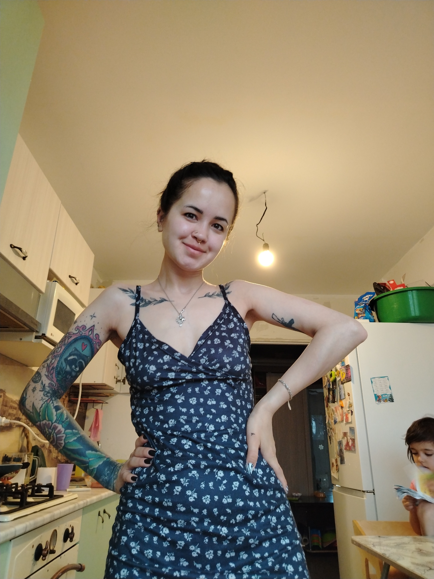 A medium, eye-level indoor shot depicts a slender, light-skinned woman standing in a kitchen with her hands on her hips. She is wearing a dark blue floral sundress with spaghetti straps and has dark hair pulled back from her face. Tattoos are visible on both arms, upper chest, and hands. The woman is smiling. In the background, a kitchen is present with white cabinets, a microwave, a stove, and a refrigerator covered with magnets and papers. A small child is sitting at a table in the lower right corner of the image, engrossed in a book or magazine. A hanging light fixture illuminates the scene. Based on provided context, the location is Noviy, Russia.