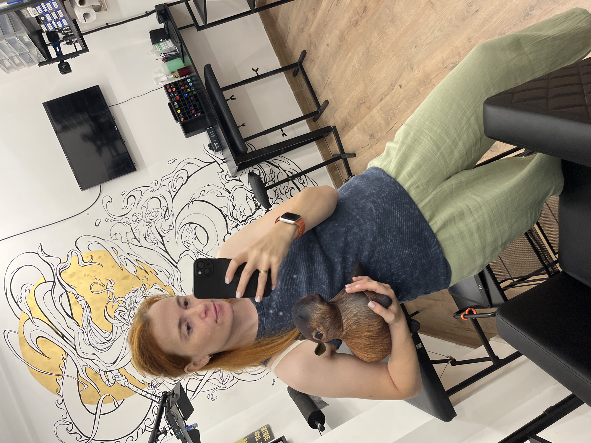 A young woman with red hair is taking a mirror selfie in what appears to be a tattoo parlor. She is holding a plush toy that resembles a capybara. She is wearing a dark blue sleeveless top and light green linen pants. In the background, there is a large mural on the wall, a television screen, and various tattoo equipment, including tattoo ink bottles, a tattoo machine, and adjustable tattoo chairs. The floor is made of light-colored wood planks. The environment appears to be a professional tattoo studio.