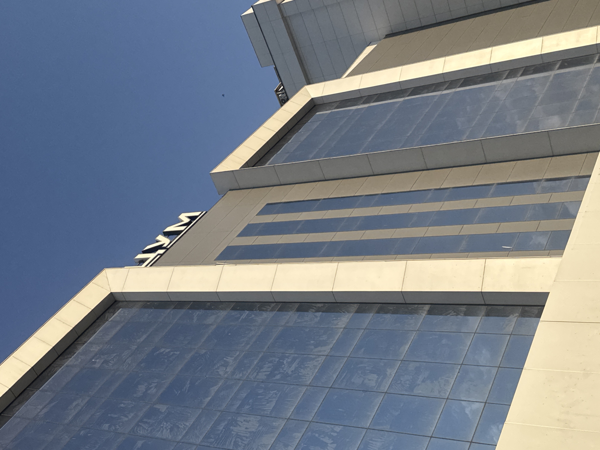 The image shows a close-up, low-angle shot of the facade of the TSUM department store in Bishkek, Kyrgyzstan. The building features large, reflective glass windows with a grid pattern, set within a light-colored, modern architectural design. The bright blue sky is visible above, with a small bird in the distance. The building's prominent signage, "ЦУМ" (TSUM in Cyrillic), is visible on the upper part of the facade. The scene appears to be an exterior view of a commercial building on a clear day.